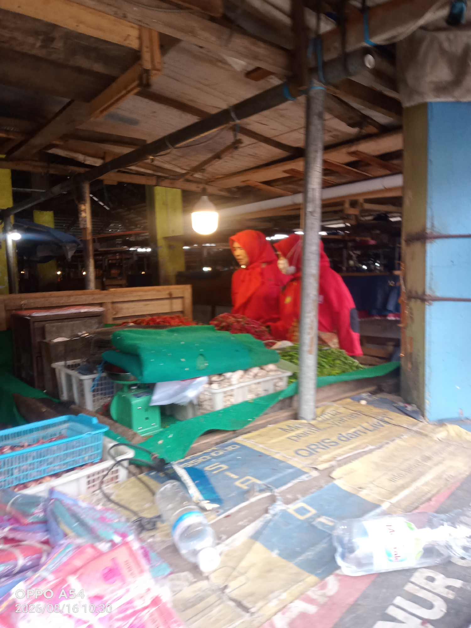 The image shows a market scene in Jambi, Indonesia. A vendor, wearing a red hijab and red clothing, is seen behind a stall. The stall displays various items, including green blankets, a green scale, and what appear to be vegetables or fruits. Crates and baskets are also visible, some containing produce and others empty. In the foreground, there are plastic bags and a water bottle on the ground. The market appears to be partially covered by a wooden roof. The overall lighting suggests it might be daytime or early evening. The specific location within Jambi cannot be determined from the image alone, but it is clearly an outdoor market setting.