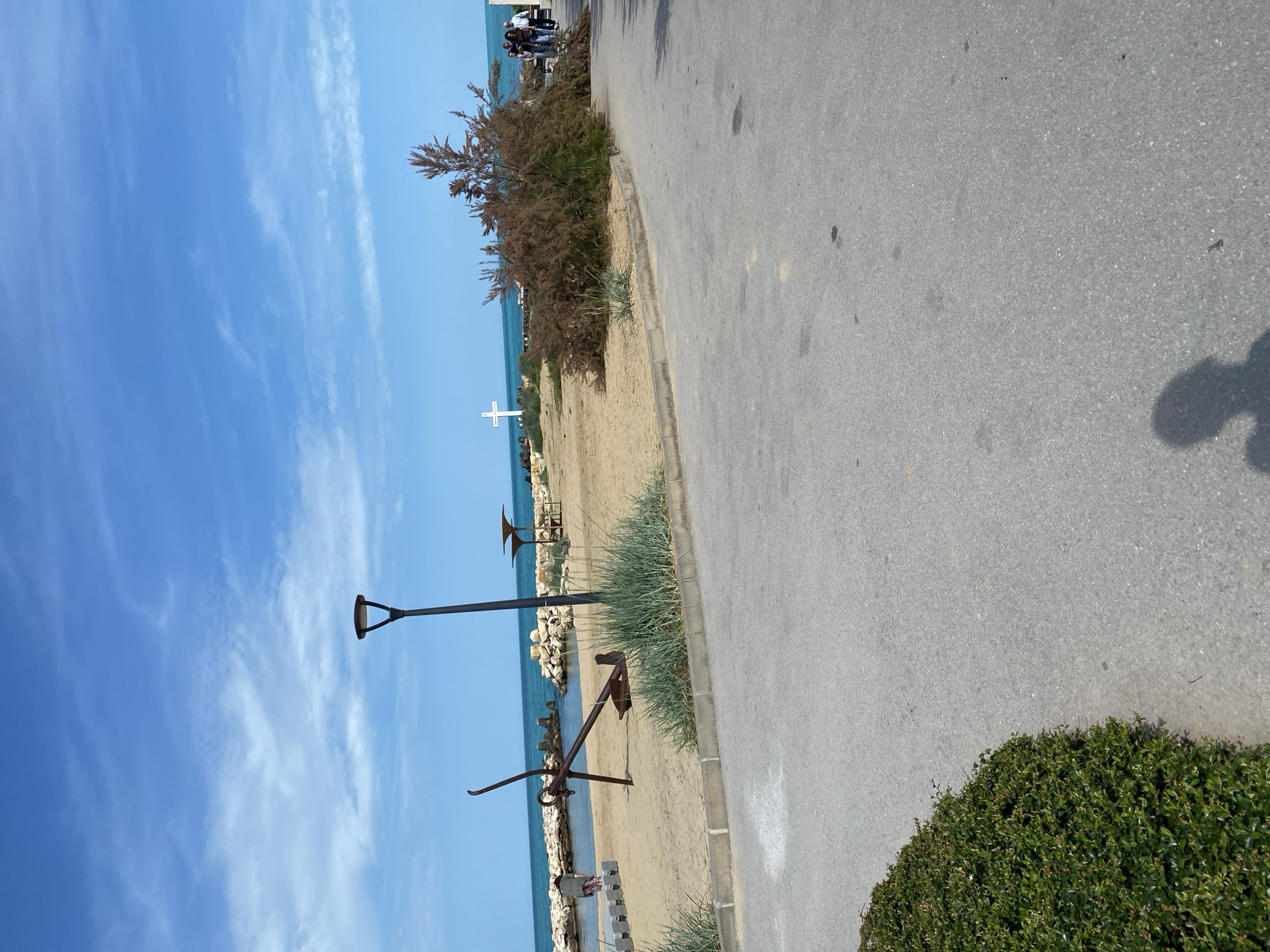 The image depicts a coastal scene with a road running alongside a beach. The road is paved and gray, with some visible dark spots and the shadow of a person. The beach is sandy and bordered by various objects, including a large rusty anchor, a white cross, street lights, and a low stone barrier partially obscured by tufts of green grass. A bush grows between the cross and the road. The sea is a vibrant blue, stretching towards the horizon. The sky is bright blue and filled with wispy clouds. In the distance, some people are visible on the beach. The image orientation is rotated. The location is Unknown, Unknown.