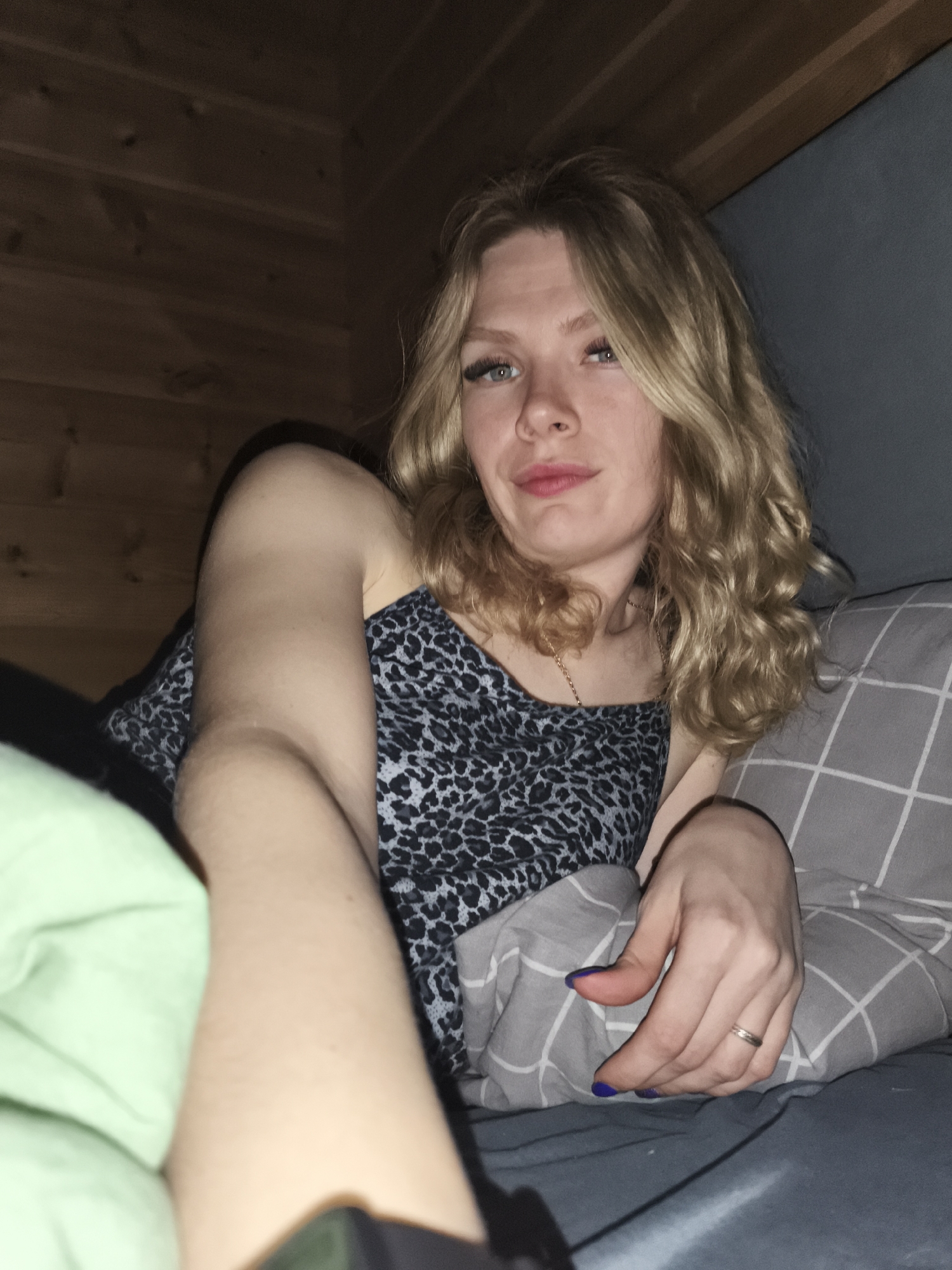 The image shows a woman lying in bed. She has blonde, wavy hair and is wearing a tank top with a leopard print pattern. She is resting her head on a gray pillow with a white grid pattern. A green blanket is visible on the left side of the frame. The background consists of wooden paneling on the left and a gray surface on the right. The image provides no information to indicate that the location is in Русский Вожой, Russia.