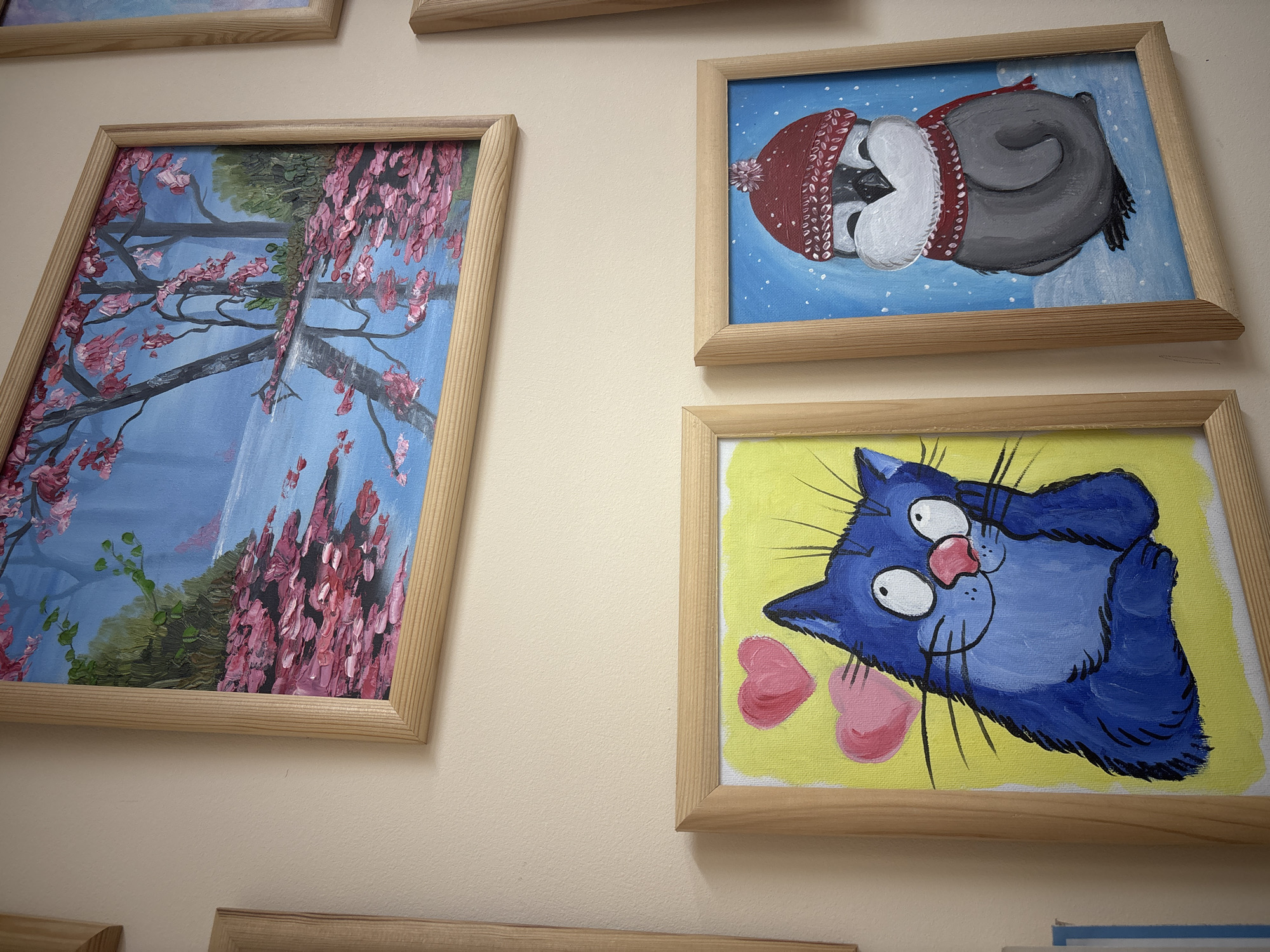 The image shows a section of a wall in what is likely an interior space. The wall is light beige. Three framed paintings are visible. The leftmost painting depicts a tree with pink blossoms against a blue background. The central painting shows a bird wearing a red hat, depicted against a blue backdrop with white dots. The third painting showcases a blue cat with pink hearts, painted on a yellow background. The location, Заволжье, Russia, is provided but cannot be confirmed or visualized within the image itself.