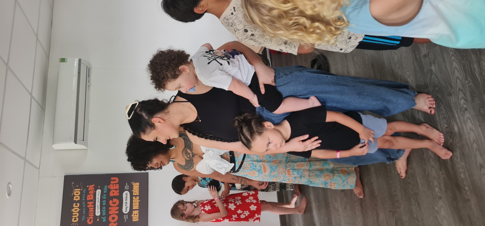 **Scene Description:**
The image shows a group of people inside what appears to be a room, possibly a studio or classroom. The group consists of adults and children, seemingly interacting or posing for a photo. An adult in a black tank top is holding several children. The background features a white wall with an air conditioning unit and a poster with text in Vietnamese.
**Location:**
The poster in the background suggests the location is likely in Đà Nẵng, Vietnam.