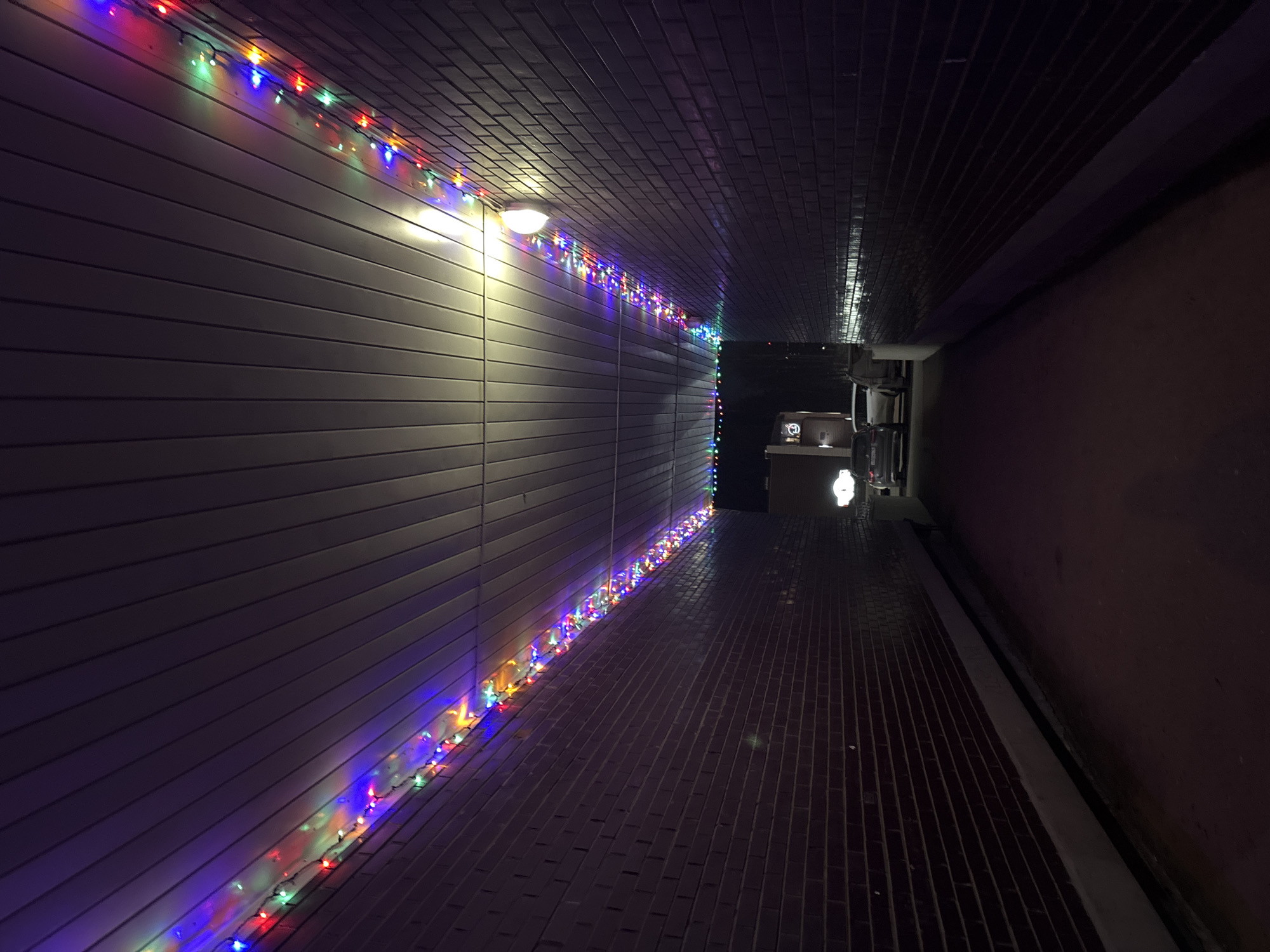 An indoor walkway with brick flooring is illuminated by a single overhead light and a string of Christmas lights on the left wall. The left wall is flat, while the right wall and ceiling are dark and textured. The end of the walkway has a dim, ambient light source, revealing equipment or objects. The location is not explicitly identifiable as Izhevsk, Russia, without further information or clues.