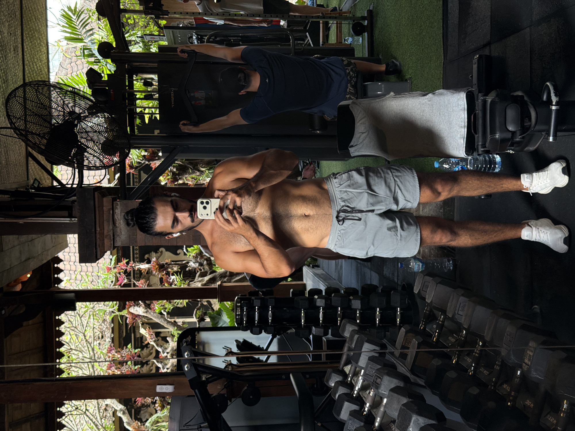 In North Kuta, Indonesia, a man with dark hair pulled into a bun is taking a mirror selfie in a gym. He is shirtless, wearing grey athletic shorts and white sneakers. He holds a smartphone in his right hand. In the background, other gym-goers are visible, one using a weight machine. The gym appears to have an open-air or tropical aesthetic, with lush greenery visible through large windows or openings in the structure. Dumbbells are neatly stacked on racks in the foreground. A large industrial fan hangs from the ceiling.