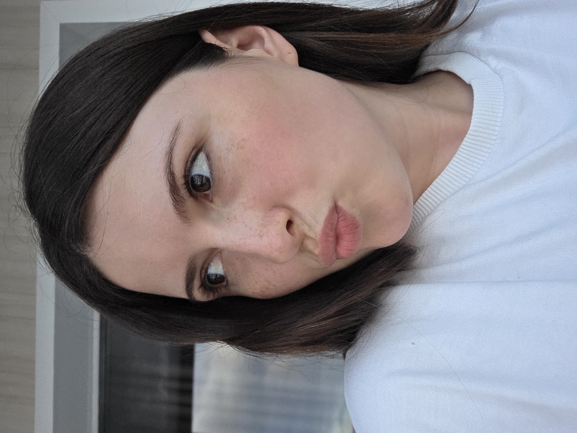 A medium shot of a young woman's face from the right side, in an outdoor setting. The woman has dark brown hair and is wearing a white sweater. She is looking to the left with her lips pursed. Her skin has light freckles across her cheeks and nose. The background is blurred but appears to be a building with windows.