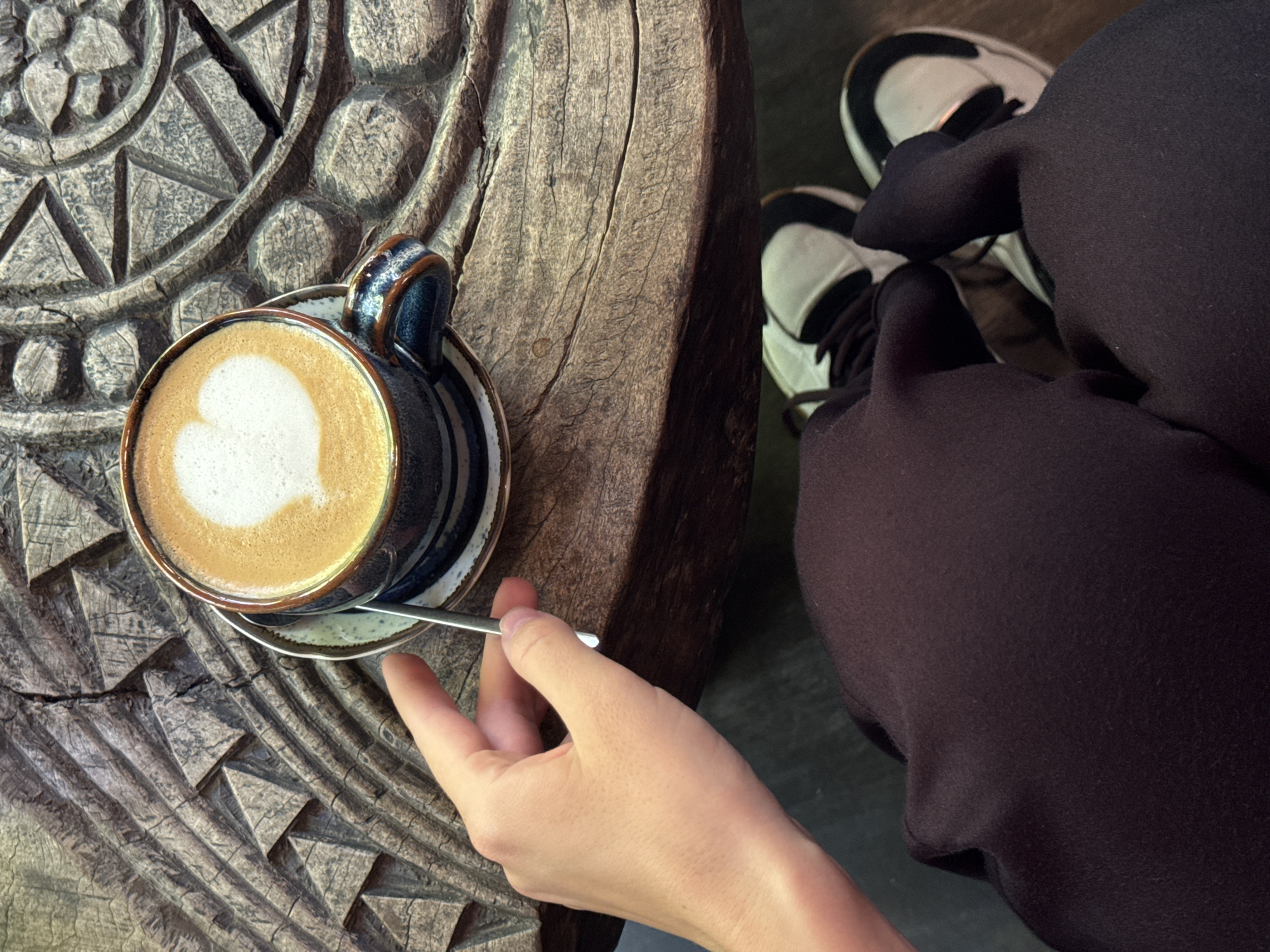 A person sits at a carved wooden table with a cup of coffee in Đà Nẵng, Vietnam. The coffee cup is blue and brown with a saucer and a silver spoon, with a heart-shaped latte art design. The person's hand is holding the spoon. The person is wearing black leggings and white sneakers with black laces. The table is intricately carved with geometric patterns. The image is taken from a high-angle perspective.