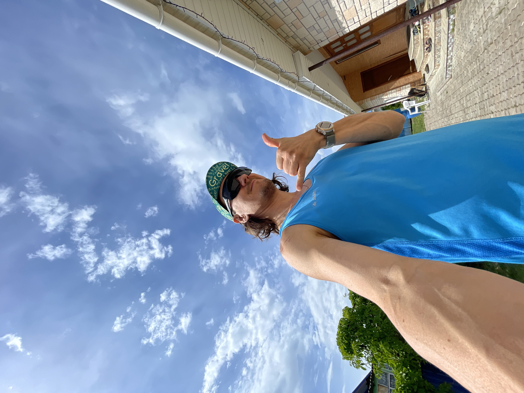 A man stands outdoors in Перевозное, Russia. He wears a blue tank top, a green cap with text, and sunglasses. He is making a "hang loose" gesture with his right hand. A wristwatch is on his left wrist. Behind him is a clear blue sky with scattered clouds. In the background is a brick building with a white gutter and a brown door. Paved ground is visible near the building.