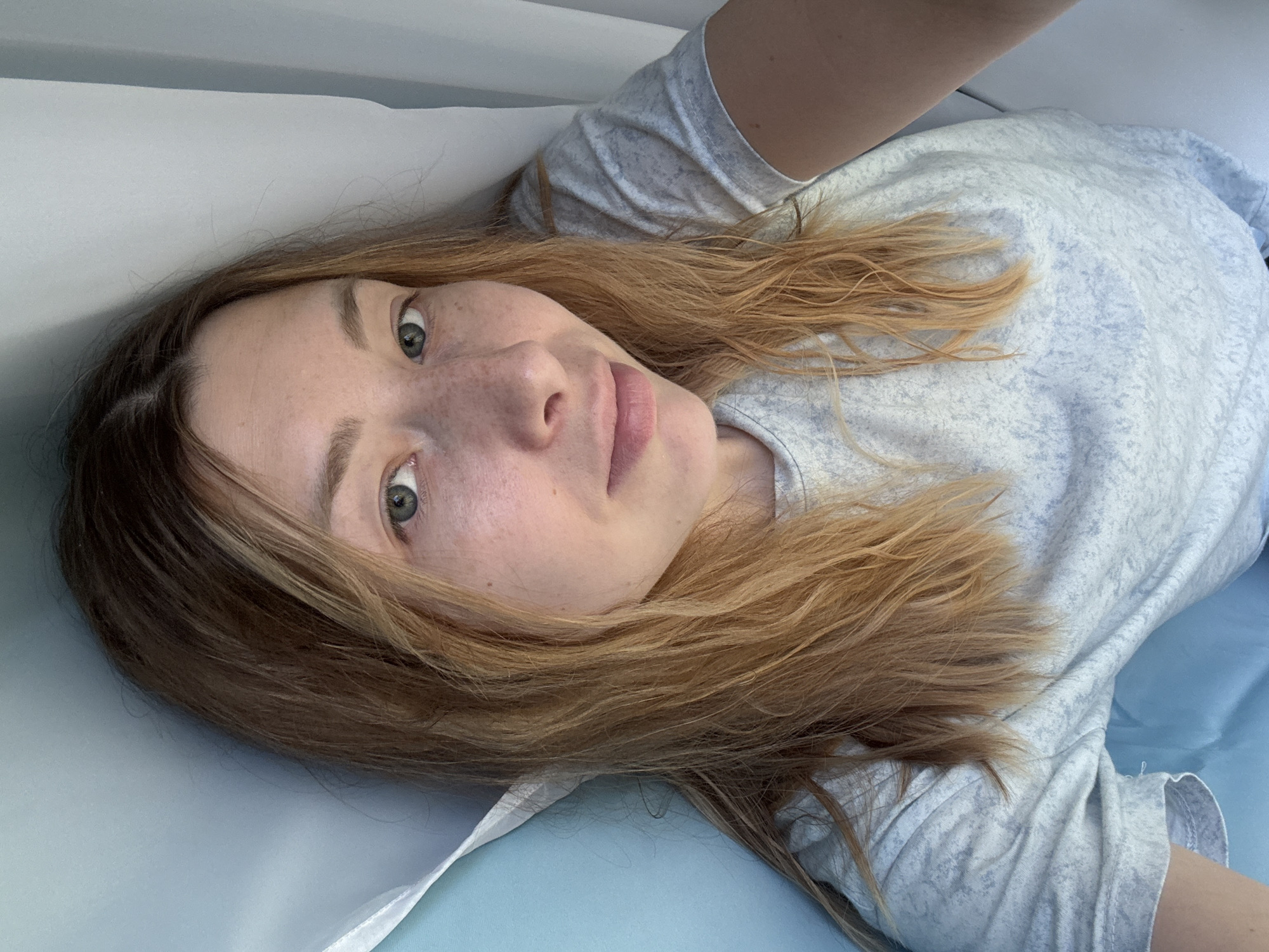 A woman with light brown hair and green eyes is lying down. Her skin has freckles. She is wearing a light gray t-shirt. The background appears to be a bed with light blue and white sheets. The location cannot be determined to be Moscow, Russia, based on the visual information provided.