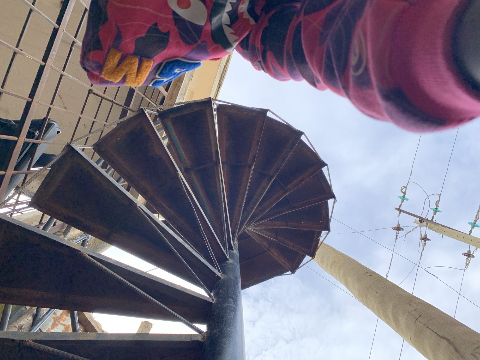 This image, captured in Rafin Par, Nigeria, presents a low-angle perspective looking upwards at a metal spiral staircase and surrounding outdoor infrastructure.
In the bottom right, a person is partially visible, wearing a distinctive hoodie. The hoodie is predominantly pink with a dark, possibly black or grey, camouflage pattern. The hood features an intricate design resembling a shark's face, including a white and red mouth area with sharp teeth, and textured yellow and blue fabric elements on the upper portion of the hood. The individual's face is not visible.
The dominant feature of the scene is a robust metal spiral staircase, occupying the left and central-right portions of the frame. It features a thick, dark, possibly black, central support pole. The individual treads are wide, dark brown or rusted metal plates, arranged in a continuous spiral. Exposed rebar is visible along the outer edges of the staircase treads, serving as structural elements.
Above the spiral staircase, to the upper right, a horizontal metal grid structure is discernible, likely the underside of a platform or ceiling. A small, light-colored fixture is attached to this overhead structure.
In the lower left and central background, a tall, light-colored wooden utility pole extends vertically into the sky. Further to the right of this wooden pole, another utility pole, appearing to be concrete, is visible. This second pole supports multiple power lines and features several sets of insulators, characterized by distinct red-brown and green-blue glass or ceramic components.
The background consists of a bright, partly cloudy sky, visible through and around the various structures.