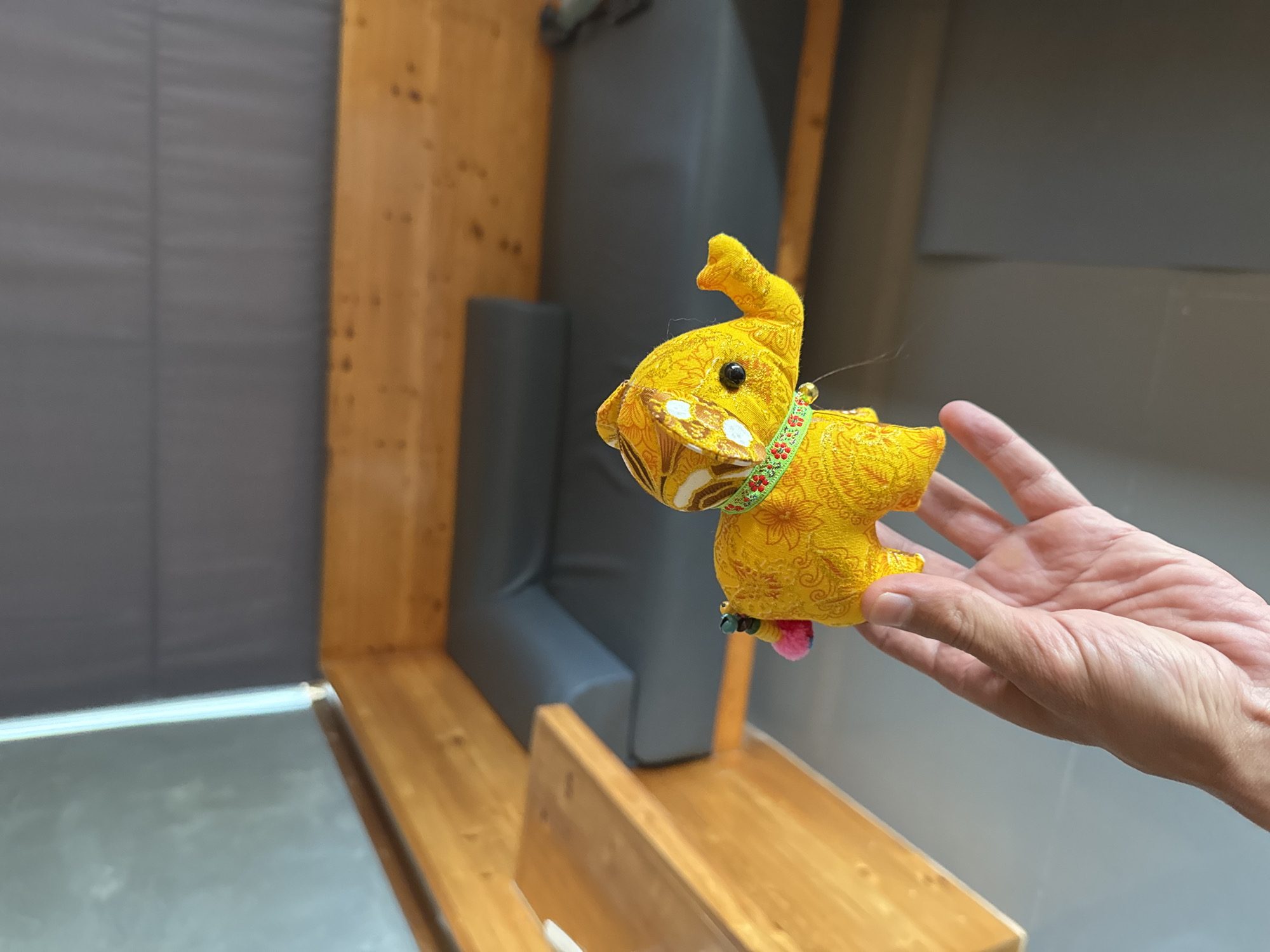 A hand holds a small, yellow, stuffed elephant charm with gold accents and a green beaded collar. The elephant has a pink pom-pom tail. In the background, a wooden bench with grey cushions is visible, suggesting an indoor seating area. The overall scene implies a casual, relaxed environment, possibly in a cafe or lounge. The image is shot from a slightly elevated angle, with the hand and elephant in the foreground.