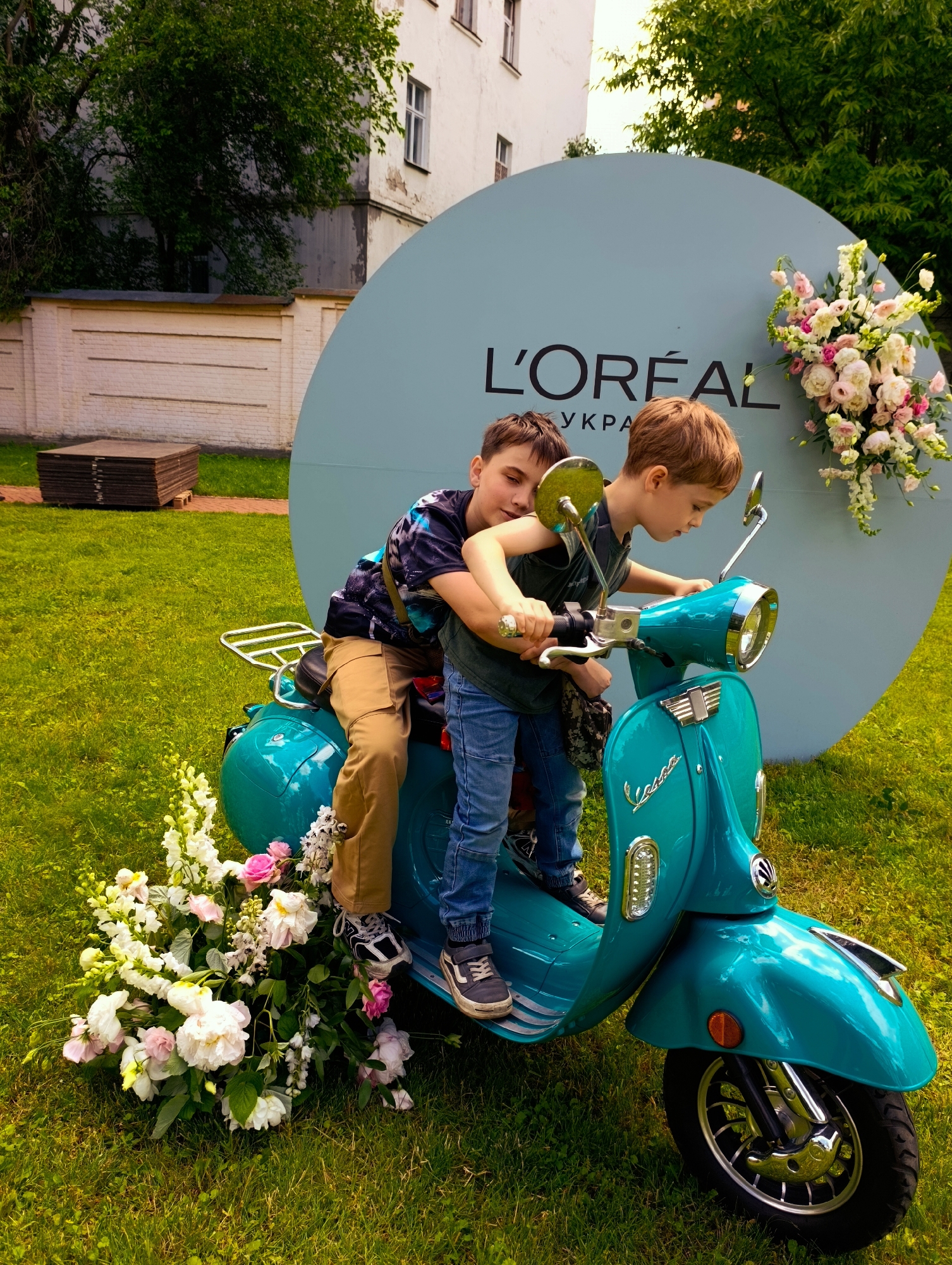 Two young boys are on a teal-colored Vespa scooter in an outdoor grassy area. The boy in front is wearing a dark t-shirt and blue jeans. The boy behind him is wearing a dark patterned t-shirt and tan pants. Both boys are looking down at the scooter. They are positioned in front of a large, light blue circular backdrop with the L'OREAL logo and "УКРА" in black text. A floral arrangement of white and pink flowers is to the right of the backdrop and another arrangement is on the left side of the scooter. A brick wall and a building are visible in the background. The overall scene appears to be a promotional event or photoshoot in Kyiv, Ukraine.