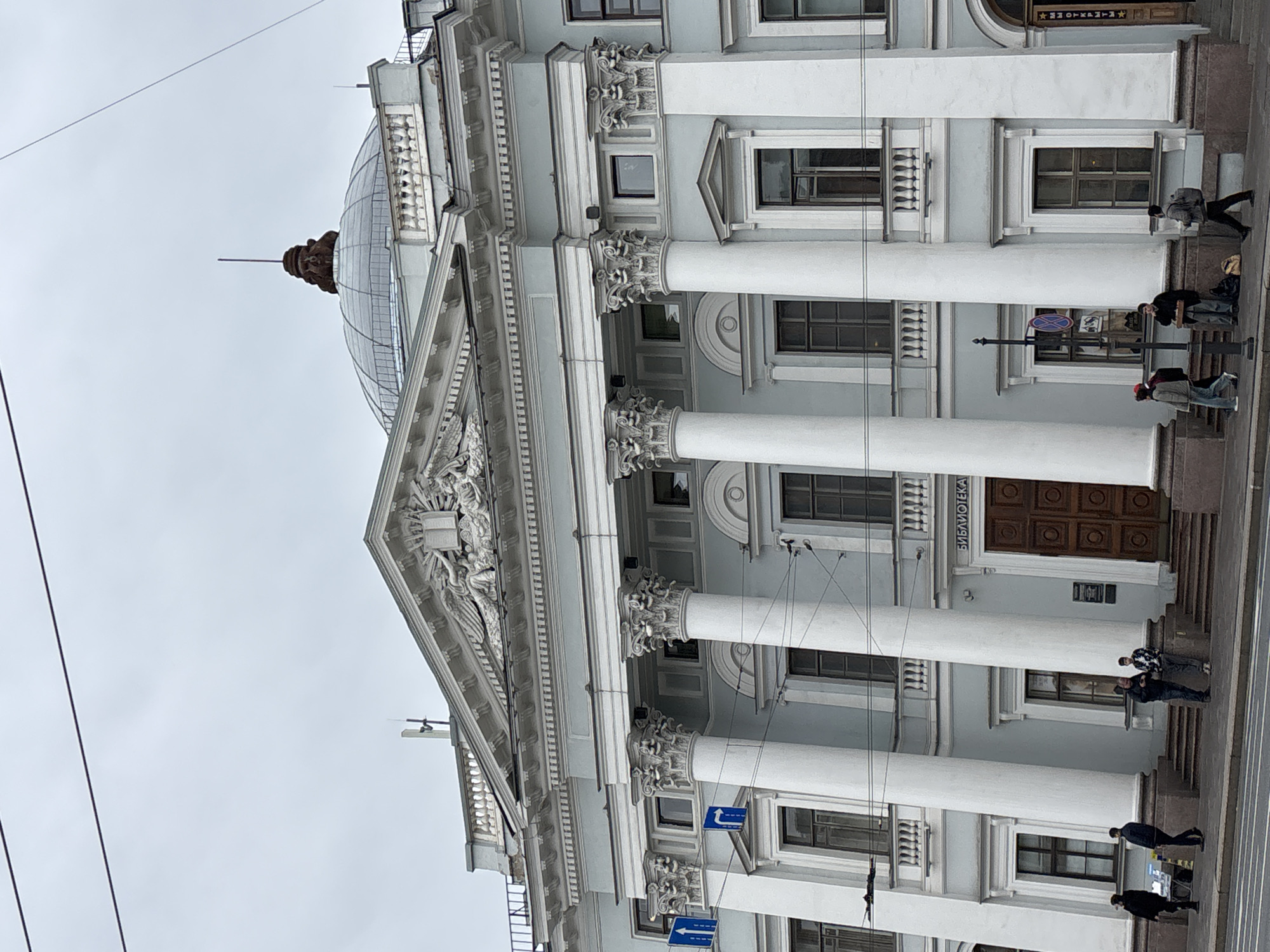 The image shows the facade of the Russian National Library building in Saint Petersburg, Russia. The building has a classical architectural style, featuring white columns, a pediment with sculptures, and a glass dome on top. The facade is light blue. The main entrance is visible, labeled in Russian with the word "Библиотека" (Library). People are walking on the sidewalk in front of the building. There are street signs present. The sky is overcast.