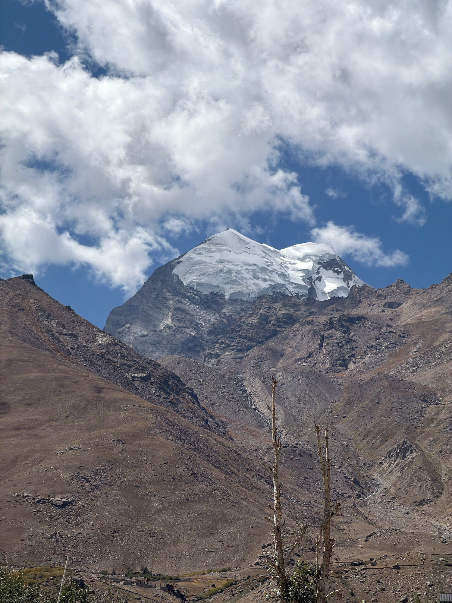 The image captures a majestic mountain range under a partly cloudy sky. The peaks are snow-capped, with rugged, rocky slopes in the foreground. The terrain appears arid and sparsely vegetated, with some dry trees visible in the middle ground. In the distance, a small cluster of buildings suggests a settlement. The scene conveys a sense of remote wilderness and natural grandeur.