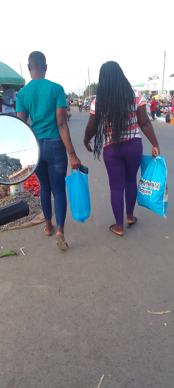Two women walk away from the viewer on a paved street, likely in a bustling market area of Kakunga, Kenya. The woman on the left has short, dark hair and wears a teal t-shirt, dark blue skinny jeans, and light-colored sandals. She carries a bright blue shopping bag and a dark smartphone in her left hand. The woman on the right has long black braided hair and wears a striped white and orange/red short-sleeved top, purple leggings, and dark sandals. She holds a bright blue shopping bag with "LONDON Shoppers" printed on it in her right hand.
In the immediate foreground, to the left, a side mirror of a vehicle (possibly a motorbike) reflects a market stall. The reflection shows piles of red produce, resembling tomatoes or fruit, and what appears to be dried fish or other market goods. Partial text "GHE..." is visible on a container within the reflection. The women are walking past this market stall.
The background reveals a street scene with numerous pedestrians, motorcycles, and various market stalls or shops lining the road. The weather appears clear and bright, indicating daytime, possibly late afternoon. The road surface is asphalt with some scattered debris like leaves and twigs. The scene captures a moment of daily life in a vibrant local street environment.