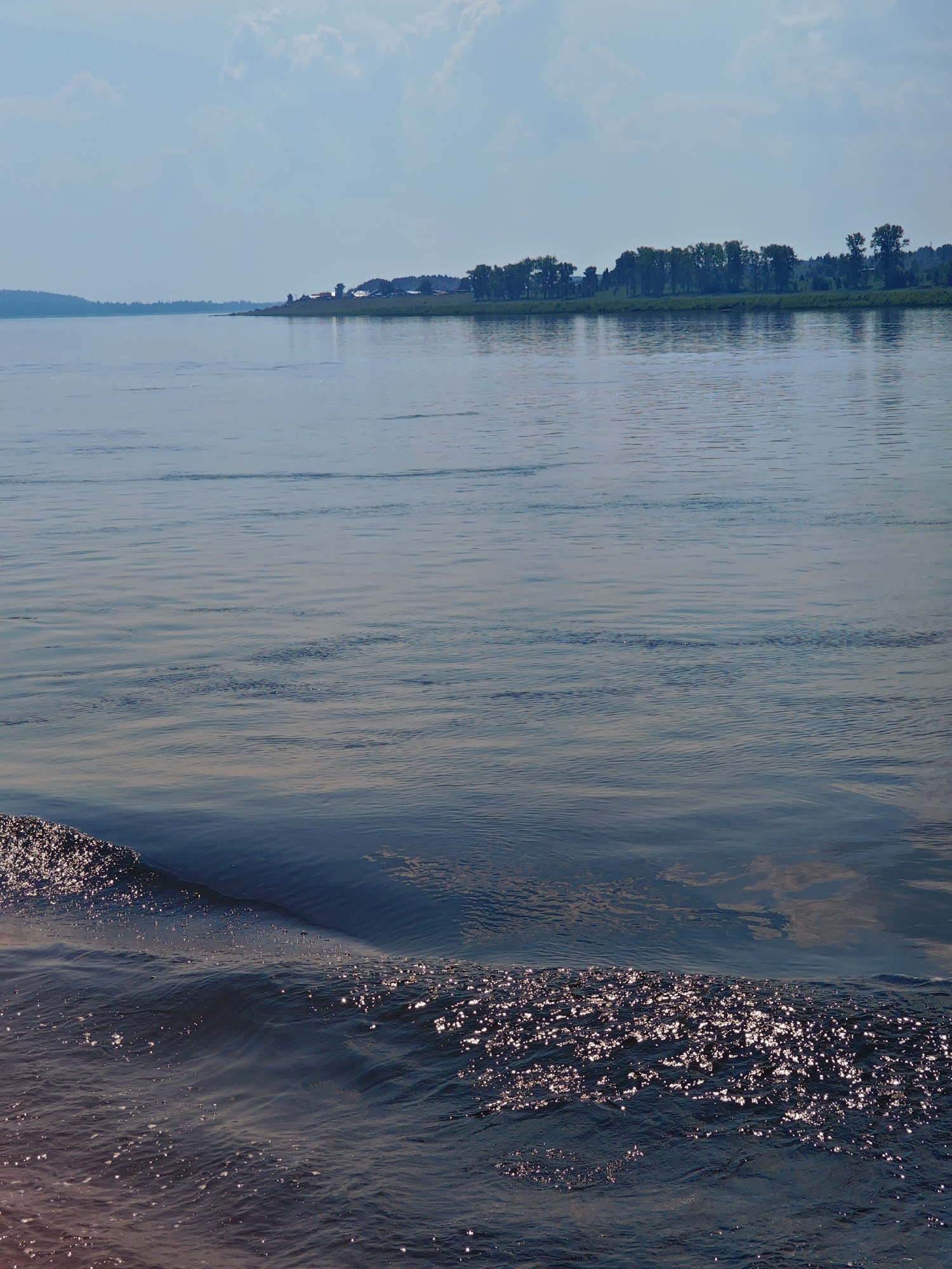 The image shows a wide expanse of calm water, likely a river or lake, with a tree-lined shore in the distance. The sky above is a pale blue with scattered clouds. In the foreground, the water exhibits gentle ripples and reflects the sunlight, creating a shimmering effect. The shore is dotted with trees and appears to have some buildings or structures visible. The overall scene is tranquil and natural, suggestive of a rural or semi-rural environment. The location is identified as Pogodaevo, Russia. There are no people or animals visible in the image.