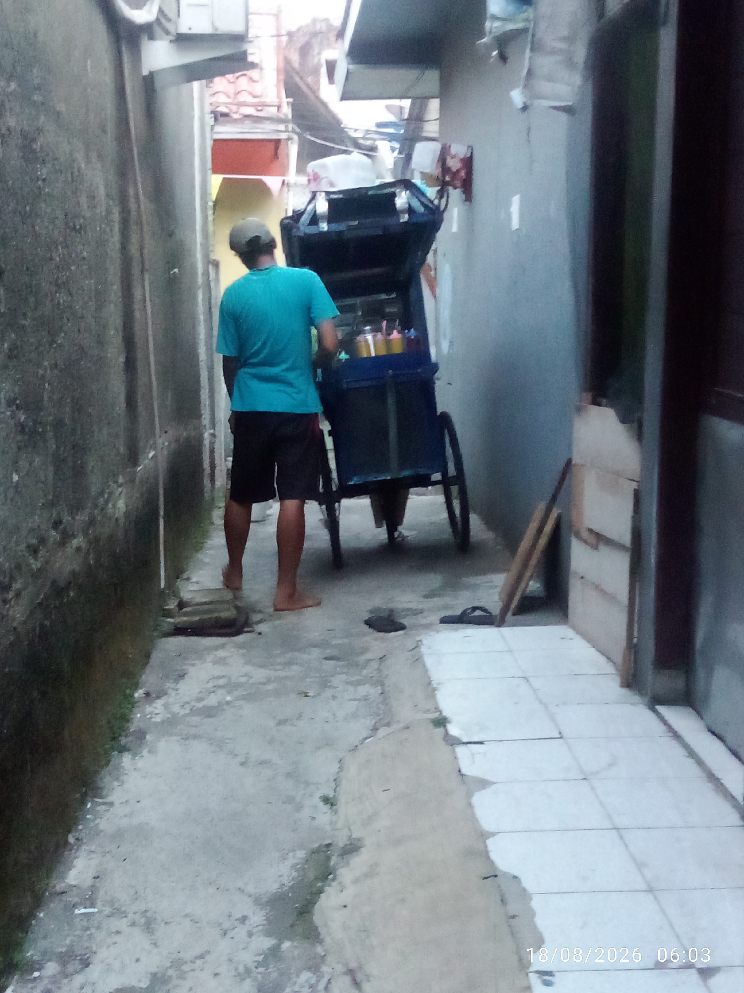 The image captures a narrow alleyway in East Jakarta, Indonesia. A man wearing a teal t-shirt and black shorts stands barefoot, facing away from the camera, looking at a blue pushcart. The pushcart appears to be a food or drink vendor, with several bottles or containers visible inside. The alley is paved with concrete, and one side features a rough, textured wall, possibly made of concrete. The other side of the alley has a lighter-colored wall, likely painted. The ground is uneven, with some debris and patches of moss. In the background, buildings and structures are visible, suggesting a residential or commercial area. A wooden plank leans against the wall on the right side of the frame, and a pair of flip-flops are discarded on the ground near the pushcart. The scene is illuminated by ambient light, and a timestamp in the lower right corner reads "18/08/2026 06:03".