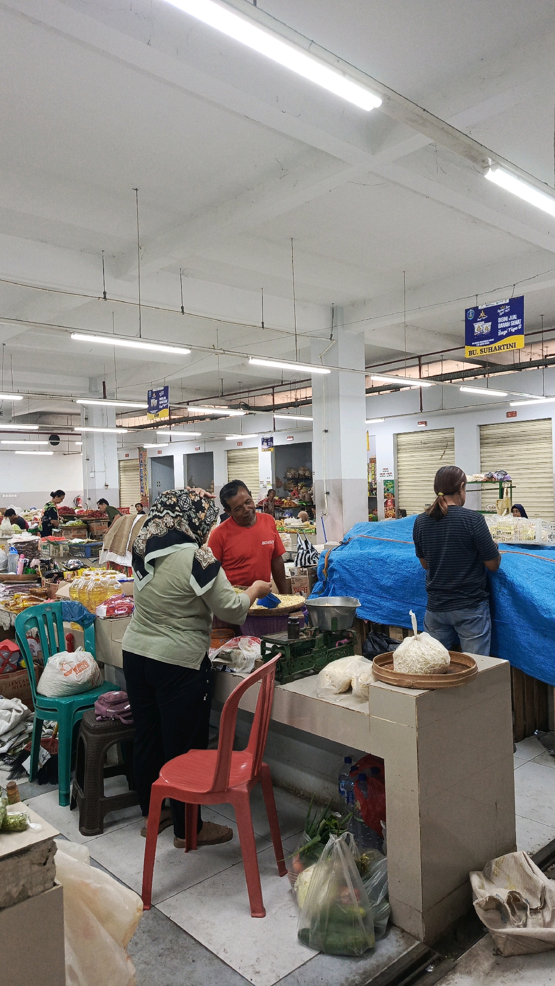 The image depicts the interior of a bustling marketplace in Ponorogo, Indonesia, likely a traditional wet market or a section of a larger indoor market. The scene is brightly lit by overhead fluorescent lights, reflecting off the white ceiling and grey concrete floor. Stalls are set up with various goods, and people are engaged in transactions.
In the foreground, a woman wearing a green hijab and a light green shirt is interacting with a man in a red t-shirt. The man is smiling and holding a scoop. They appear to be at a stall where dry goods, possibly corn or grains, are being sold. In front of them is a weighing scale and containers of food items.
To the left of the main interaction, there are plastic chairs, one red and one teal, and various bags and containers. Bottles of cooking oil are visible on a shelf. Further back, other shoppers and vendors can be seen amidst displays of produce and other market goods.
To the right, a woman with her hair in a ponytail, wearing a striped t-shirt and denim shorts, is standing near a large blue tarp covering some goods. Bags of produce, including onions and leafy greens, are visible in plastic bags on the floor.
Banners with Indonesian text are hung from the ceiling. One prominent banner reads "DISINI JUGA DAGANG SEGI TIGA G" and "BU. SUHARTINI," suggesting a specific stall or vendor. The architecture suggests an indoor market structure with metal beams and roll-up doors visible in the background, indicating closed stalls or shops. The overall atmosphere is one of active commerce and everyday life in a local Indonesian market.