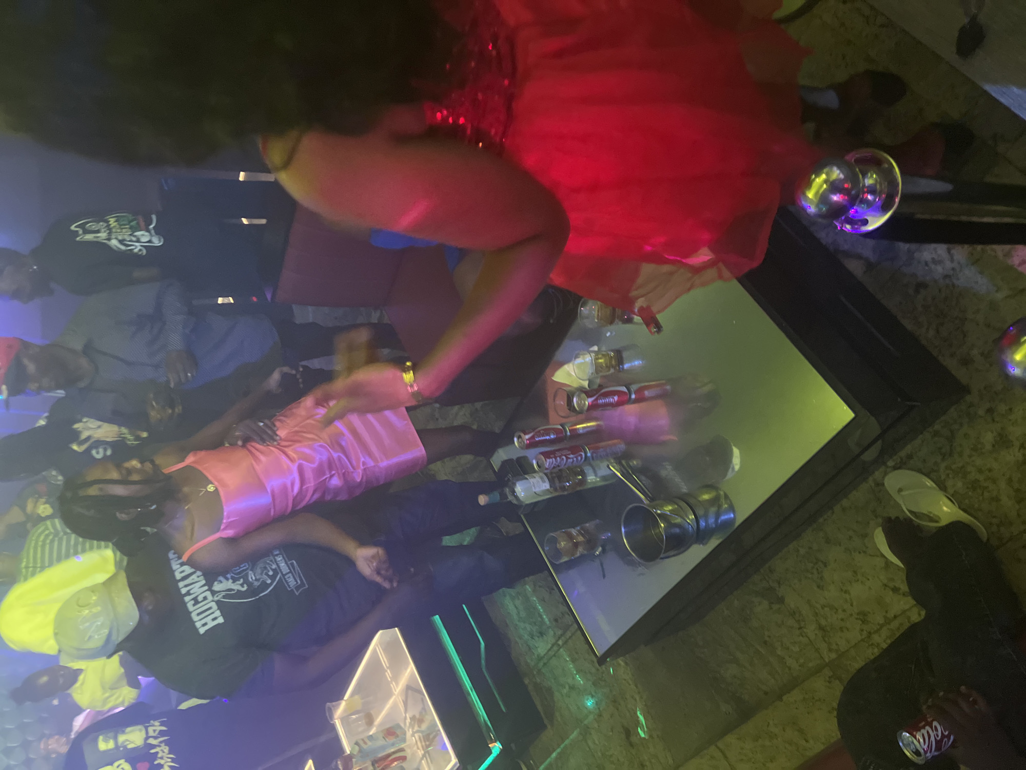 A dimly lit indoor space contains several people around a glossy, reflective table with beverages. One woman wears a red dress and another a pink dress. A man in a black shirt with "Hogwarts" printed on it is partially visible. There are cans of Coca-Cola, glasses, and a metallic object on the table. The location is unidentified, and there is no confirmation it is in Howayi, Nigeria.