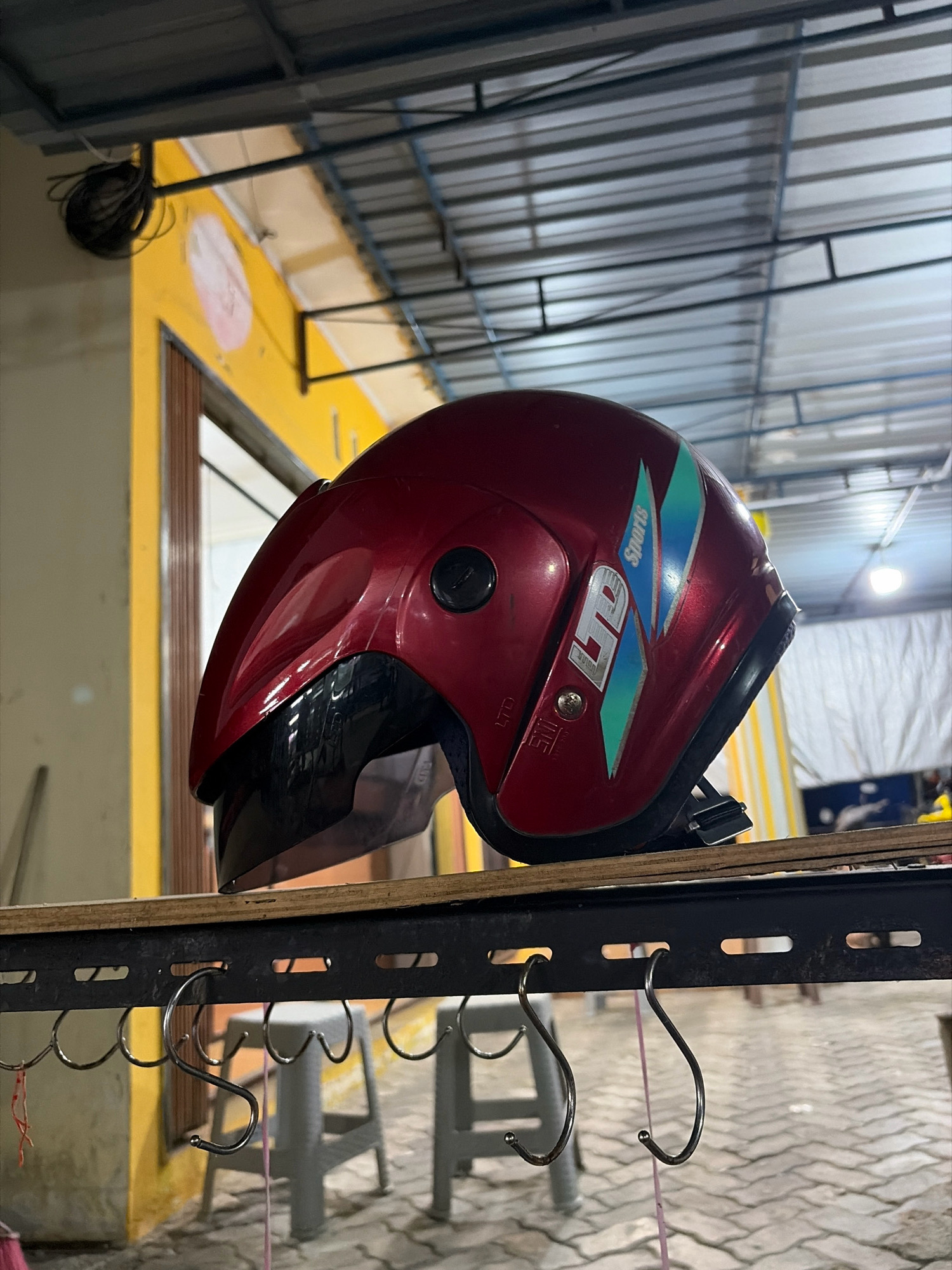 A red motorcycle helmet is displayed on a metal rack in what appears to be an outdoor market or shop in Tanjung Pinang, Indonesia. The helmet, with a dark visor and teal and white accents on the side, is positioned in the foreground. Behind it, a bright yellow building with a corrugated metal roof is visible, and a coiled black cable hangs from the eaves. The ground is paved with interlocking bricks. Two grey plastic stools are also visible on the brick surface. The overall setting suggests a commercial area, possibly a roadside stall or shop, common in Indonesian cities like Tanjung Pinang.