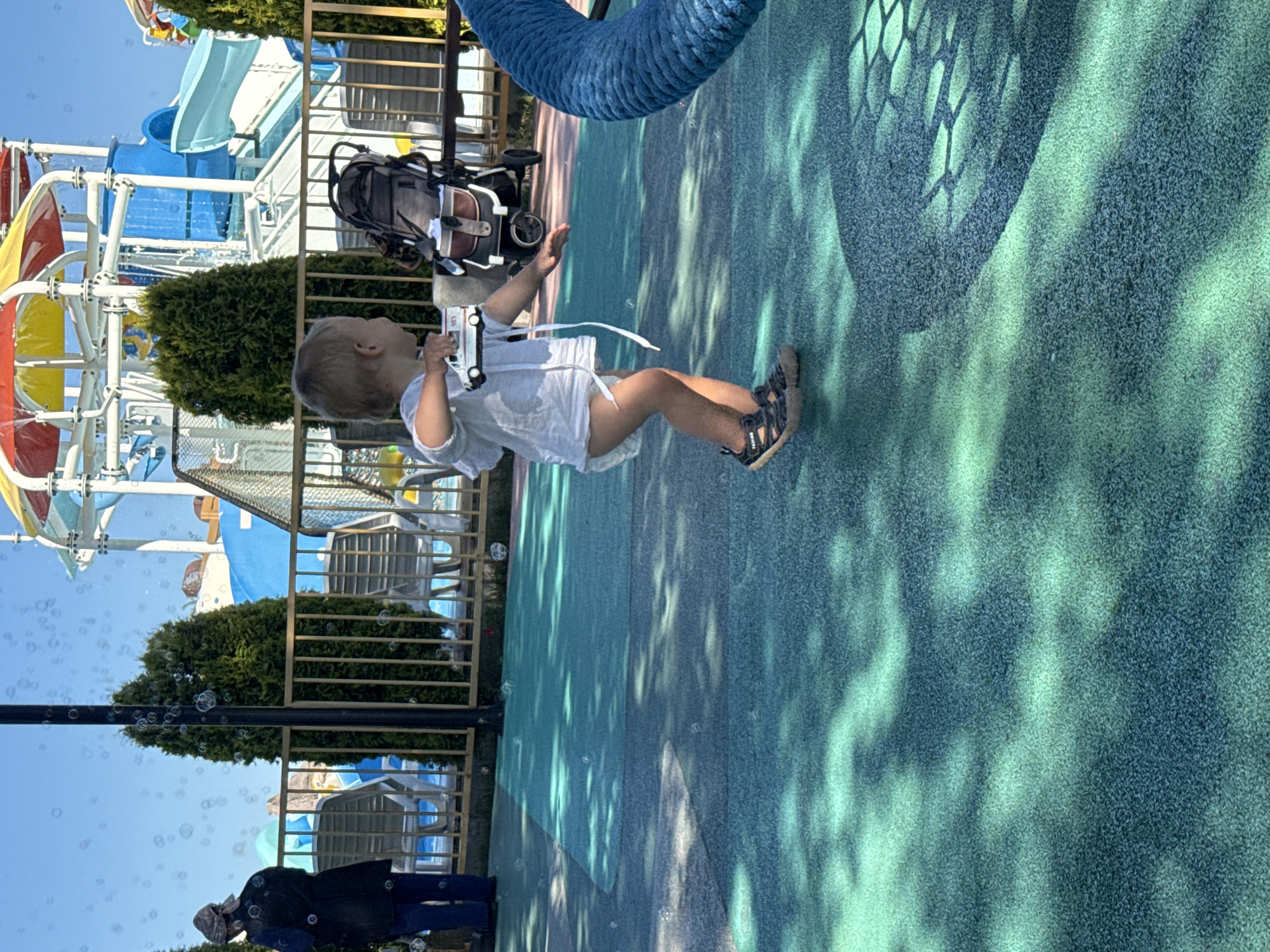 The image shows a young child playing with a toy car on a patterned, rubberized surface. The child is wearing a light-colored shirt, a diaper, and sandals. In the background, there are trees, lounge chairs, a fence, and part of a water park with slides. A stroller is visible near the child. The scene is outdoors, with a clear blue sky and bubbles floating in the air, suggesting a sunny day. This location is a playground area at a water park in Sochi, Russia.