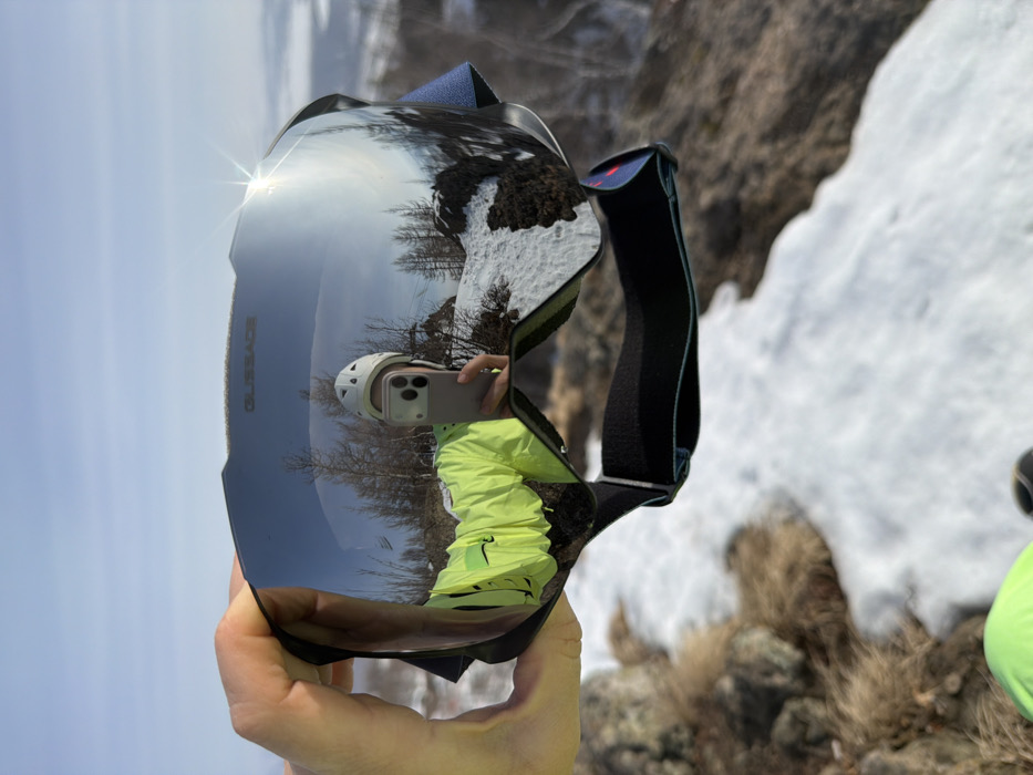 A right hand holds a pair of "GLISSADE" branded ski goggles with dark, reflective lenses, centered in the frame. The goggles' surface reflects a person wearing a white helmet and a bright neon yellow/green jacket, holding a light-colored smartphone to capture a self-portrait. This reflection also displays a clear blue sky, bare deciduous trees, and snow-covered rocky terrain. A prominent sun flare is visible on the upper right portion of the goggle lens. The background directly visible beyond the goggles features a landscape of white snow patches interspersed with dry, light brown grass, exposed rocks, and sparse, bare trees under a pale blue sky. The scene is located outdoors in an elevated, possibly mountainous or hilly environment within Zelenaya Polyana, Russia.