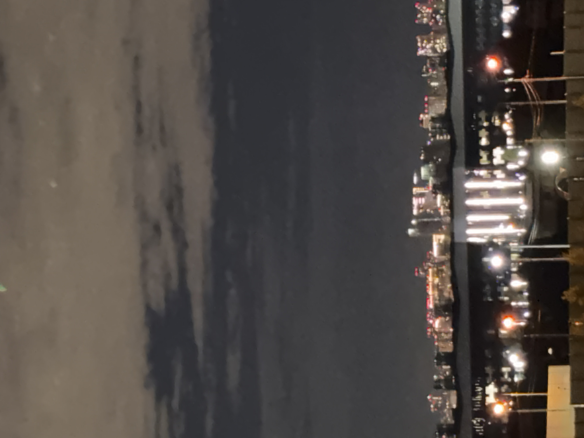 The image captures a nighttime cityscape with a focus on illuminated buildings and streetlights. In the foreground, the silhouettes of several streetlights are visible, some emitting a warm orange glow, while others are a brighter white. Behind these, a line of more distant buildings is illuminated, showcasing a variety of window lights and architectural features. The background is dominated by a dark, cloudy sky, with the lights from the city reflecting faintly off the cloud cover. The overall impression is one of urban activity at night. The image does not contain any people or specific landmarks that would identify the city as Odintsovo, Russia.