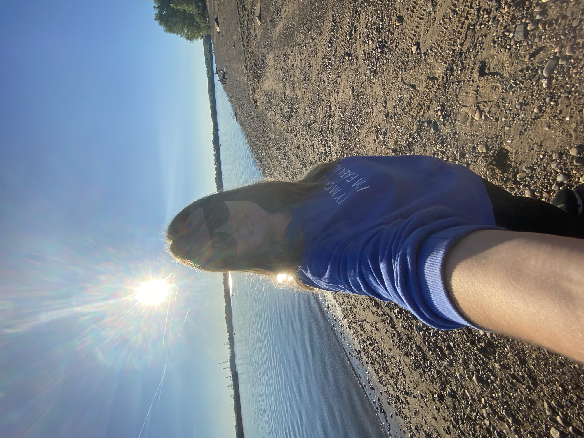 A woman is taking a selfie on a pebble beach next to a body of water. She is wearing a blue sweatshirt with white text that reads "MY MOM IS FABULOUS". She is also wearing sunglasses and has long brown hair. The sun is shining brightly and there are some clouds in the sky. In the background, there are some boats on the water and trees on the shore. The location is believed to be in Chaikovsky, Russia, specifically along the Kama River, as indicated by the industrial structures visible in the distance, which are characteristic of the city's port.