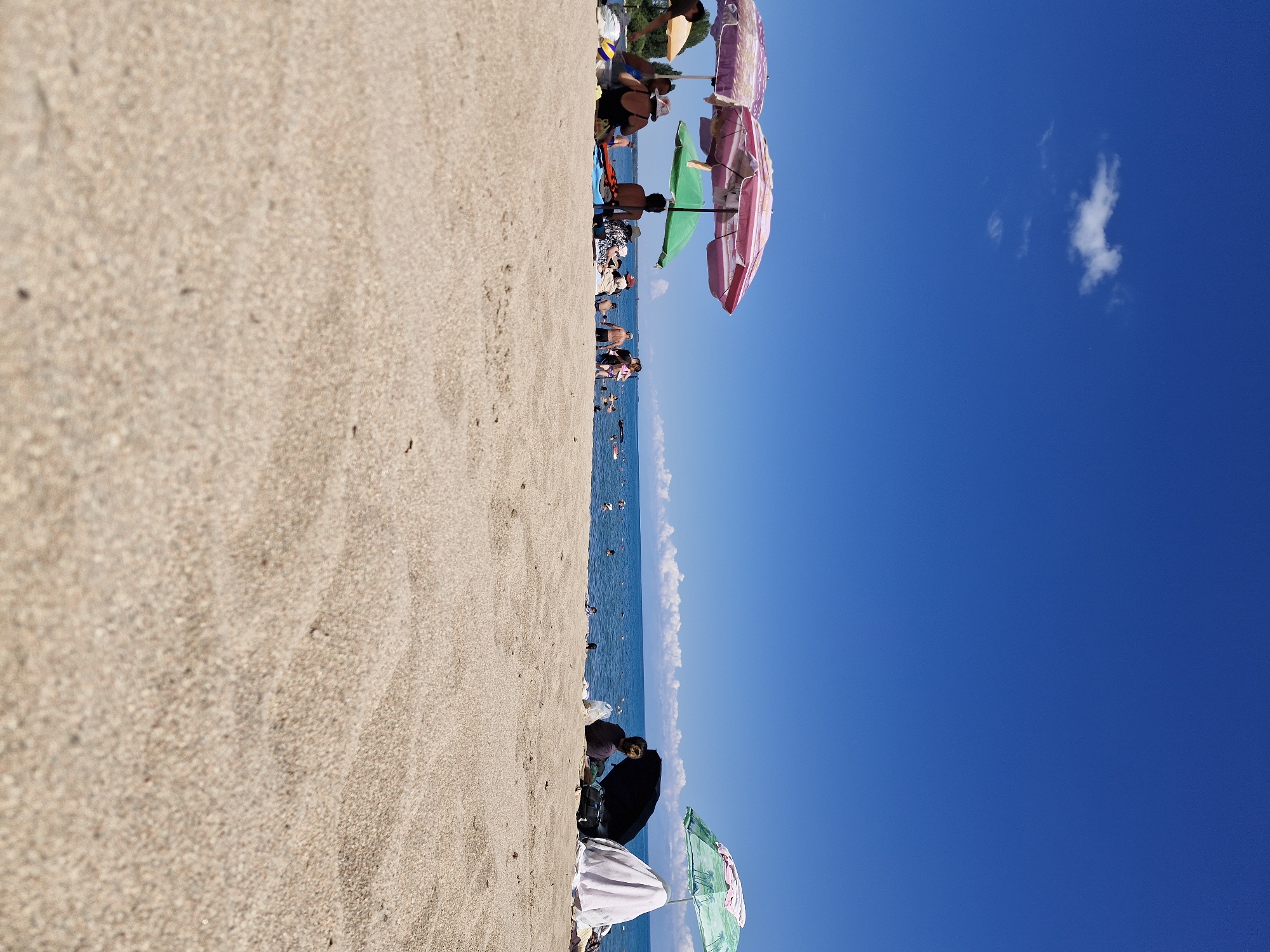A wide-angle, low-angle shot captures a bright, sunny day at a beach, likely in Bulan-Segettu, Kyrgyzstan, given the context. The foreground is dominated by sand, with the texture of grains clearly visible. In the mid-ground, a group of people are scattered across the sand, many under the shade of beach umbrellas. Several umbrellas are visible, including two with pink and purple patterns and one green umbrella. People are sitting on the sand, some appear to be relaxing or chatting. Further in the background, the calm, blue water of a lake stretches to the horizon, where a few clouds dot the otherwise clear sky. Other beachgoers are visible in the water, swimming or enjoying the lake. The scene conveys a sense of leisure and a typical summer day at a popular beach destination.