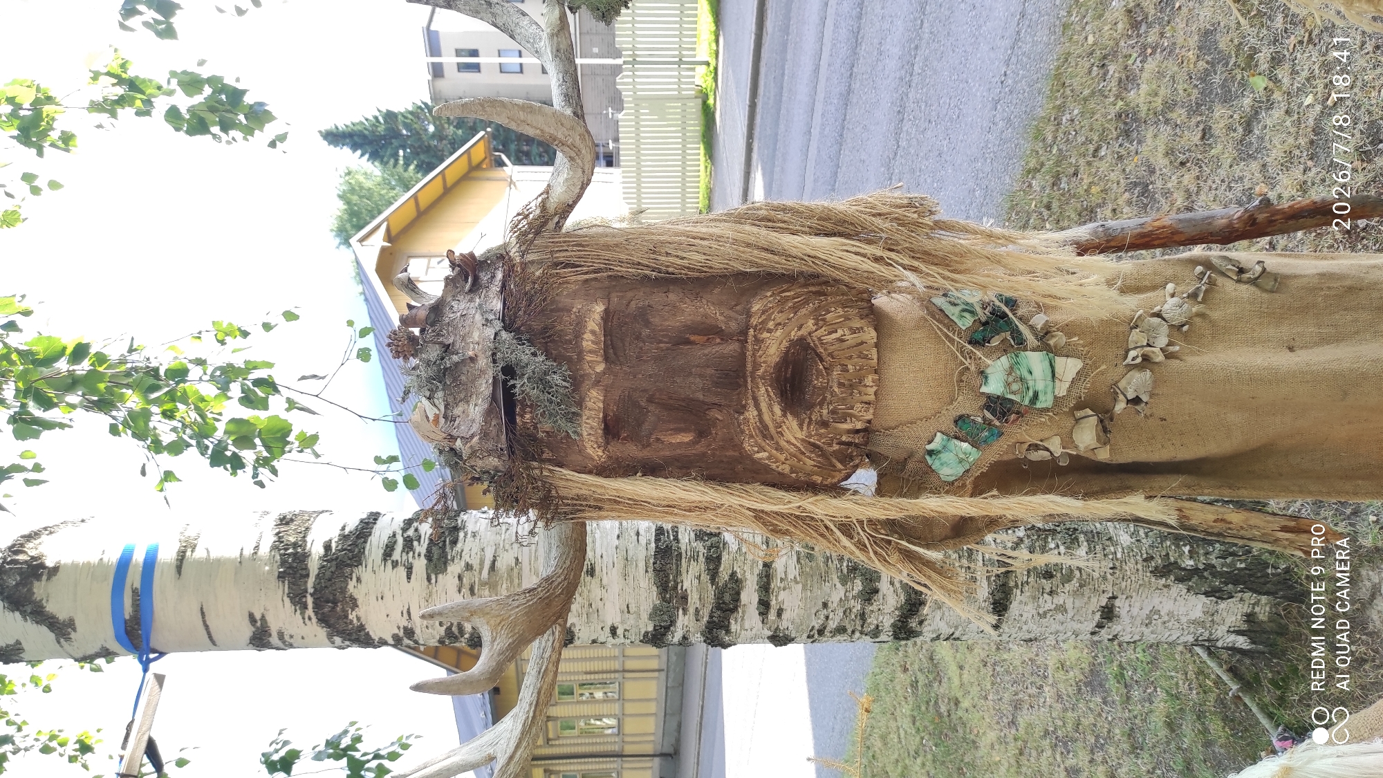 The image shows a close-up of a folk art figure made of wood and natural materials, positioned outdoors in a city setting. The figure is designed to resemble a human face with a beard and long, straw-like hair. It is adorned with large antlers, a headdress made of bark and moss, and a necklace composed of green stones and shells. The figure is dressed in a burlap-like material, with additional natural elements hanging from its body.
Behind the figure, a birch tree is visible, with a blue ribbon tied around its trunk. In the background, a yellow house with a gabled roof can be seen, along with a white picket fence and a paved road. The presence of buildings and a road indicates an urban or suburban environment.
The image also contains a timestamp and text overlay, "REDMI NOTE 9 PRO AI QUAD CAMERA 2026/7/8 18:41". This suggests the photo was taken with a Redmi Note 9 Pro phone on July 8, 2026, at 6:41 PM. While the exact location in Iisalmi, Finland, cannot be precisely determined from the visual information alone, the style of the house and the overall atmosphere are consistent with Finnish scenery. The folk art nature of the figure might suggest it is part of a local festival, exhibition, or a permanent installation in a public space.