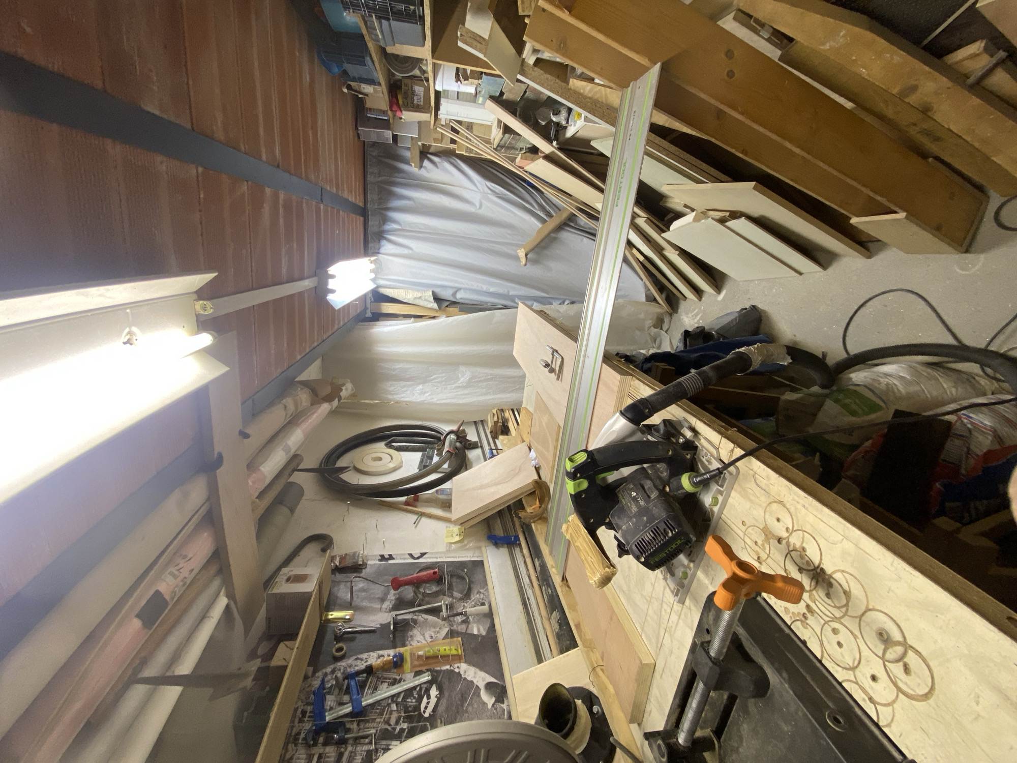 This image depicts an indoor workshop or storage space, appearing to be in an active state of use for woodworking. No specific visual cues within the image indicate its location in the city of Paris, France.
The ceiling features exposed terracotta-colored tiles interspersed with dark, horizontal structural beams. Illumination is provided by two long, fluorescent strip light fixtures mounted to the ceiling.
In the foreground and central-left, a light-colored wooden workbench dominates the scene. The workbench surface displays a pattern of overlapping circular burn or cut marks. An orange, three-pronged clamp handle extends from a dark metal mechanism on the workbench. Positioned on the workbench is a black and green Festool track saw, with a black corrugated dust extraction hose attached. A long, silver Festool guide rail extends diagonally across the workbench from the saw. Several pieces of light-colored plywood and other wood stock are arranged on the workbench and to its left. Part of a larger, dark-colored stationary saw (possibly a miter saw) is visible on the far left.
The wall behind the workbench, to the left, is adorned with a large, black and white poster depicting an urban street scene. Various tools, including clamps, a spirit level, and a red-handled implement, are hung on the wall. A coiled black hose or cable hangs to the right of the poster.
Towards the rear and right of the workshop, a large grey tarp or curtain hangs, possibly concealing a storage area or doorway. Numerous wooden planks and sheets of various sizes are stored vertically and horizontally on the floor against the back wall, forming a wood-stock pile. Shelving units with assorted boxes and containers are visible on the far right. The floor is a light-colored concrete or similar material. No individuals are present in the scene, and no active work or interactions are depicted.