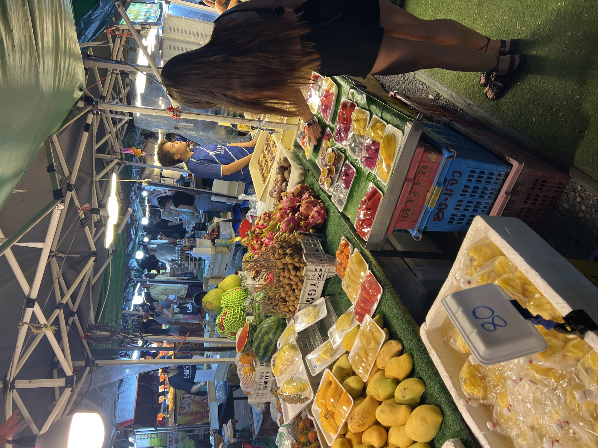This image captures a bustling fruit stand at a night market in Baan Bo Phut, Thailand.
A woman with long brown hair and wearing a dark dress and sandals is standing at the fruit stand, seemingly examining the pre-cut fruit platters on display. The vendor, a middle-aged man in a blue shirt, stands behind the counter. He is looking at the woman.
The fruit stand is filled with an abundance of tropical fruits, including mangoes (both whole and pre-cut), dragon fruit, pomelo, rambutan, watermelon, durian, and longan. They are arranged in a vibrant, colorful display.
The lighting is a mix of natural and artificial. It appears to be under the cover of a tent. In the background, other stalls and people can be seen, indicating a lively market atmosphere.