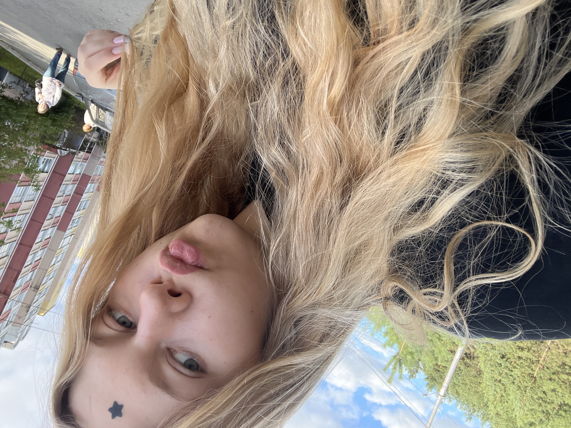 A blonde woman with light blue eyes and a dark star sticker on her forehead takes a selfie outdoors. She is wearing a black top and her long blonde hair is partially obscuring her face. Her lips are pursed. In the background, a multi-story building with red and yellow accents is visible, along with trees and a cloudy blue sky. Two other women are in the distance, one standing near a car and the other walking on a paved path. The scene appears to be a residential area in the city of Perm, Russia.