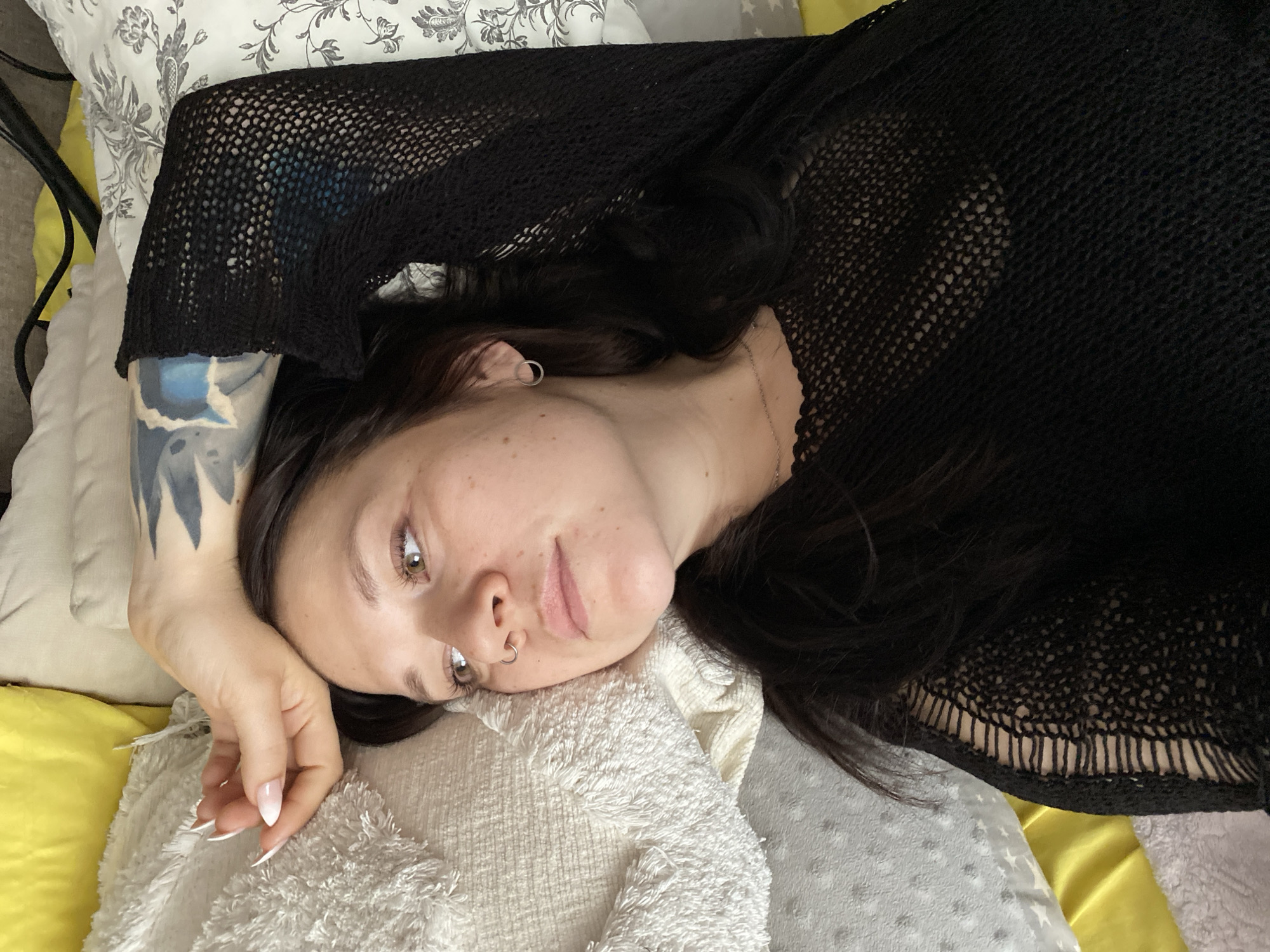 A woman with fair skin, dark hair, and light brown eyes is lying on a bed with multiple pillows. She wears a black, crocheted-like top, a silver chain necklace, and a silver nose ring. A tattoo is visible on her left forearm. Her long nails are manicured with a light pink to white gradient. The pillows are various colors and textures, including white patterned, yellow, white furry, and gray with stars. The location is not identifiable as Chaikovsky, Russia from the given image.