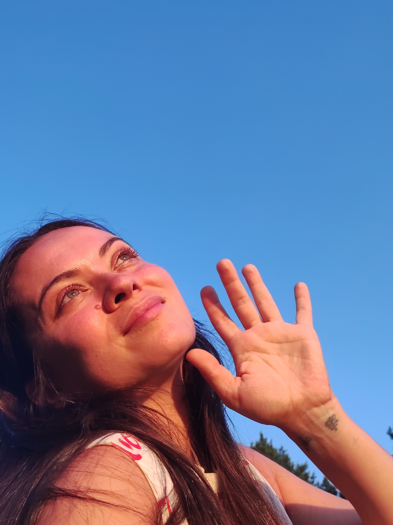 This is a medium shot, low-angle, outdoor photograph of a young woman looking up at a clear blue sky. The sun is shining brightly, casting a warm glow on her face and creating a slight shadow from her raised hand. Her expression appears serene and hopeful as she gazes upwards. Her hand is held up, palm facing the camera, partially obscuring her face. A small tattoo is visible on her wrist. She has long, dark hair that frames her face. She is wearing a white top with red lettering. The background is dominated by the vast expanse of the blue sky, with the tops of some evergreen trees visible in the lower right corner. The lighting suggests it is either early morning or late afternoon, with the sun low on the horizon. The image evokes a sense of peace and connection with nature.