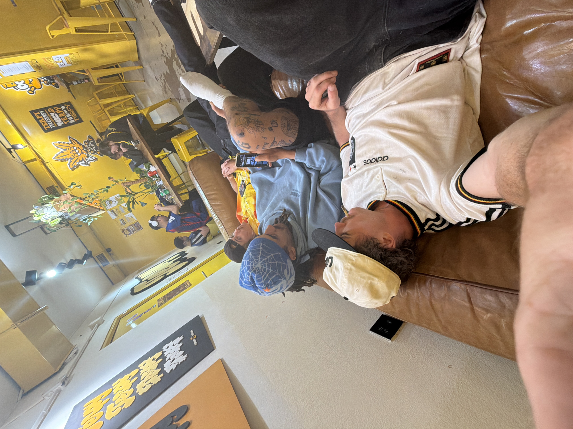 A group of four young adults is reclining on a brown leather couch inside what appears to be a restaurant or bar setting. One person wears a white Adidas jersey and a beige baseball cap, while another has a blue beanie with a white spider web design. The person next to them is looking at their phone, and another has tattoos on their head. The environment is characterized by yellow walls, yellow furniture, and various posters and signage in Asian characters. This location, based on the distinctive decor and signage, is likely a business named "Terpy" in Barcelona, Spain.
