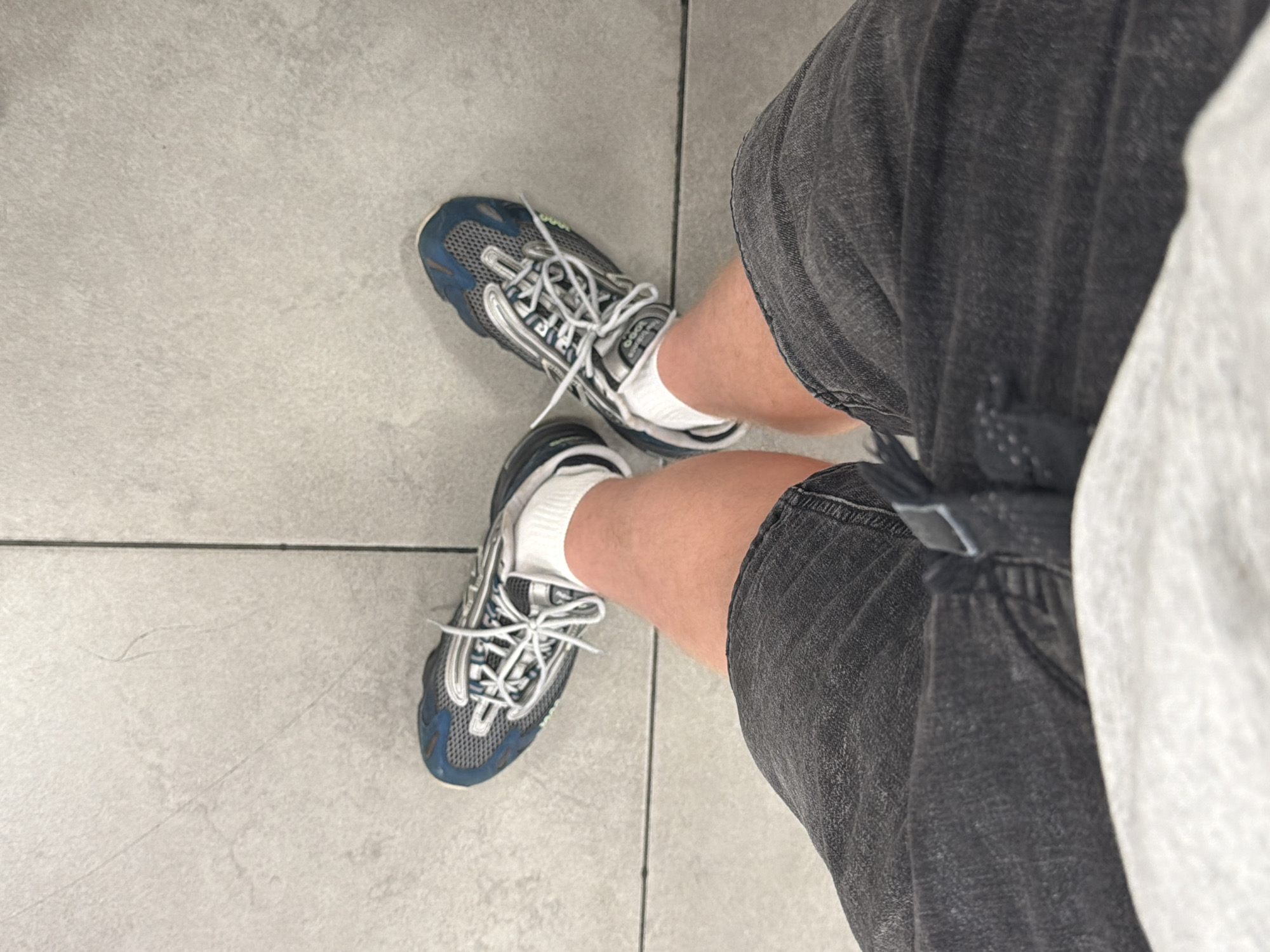 A first-person perspective shot shows a person standing on a light gray tiled floor. The person is wearing dark gray denim shorts and white socks. On their feet are blue and silver sneakers with white laces. The image is taken from above, looking down at the person's feet and lower legs. The location is Niles, United States of America.