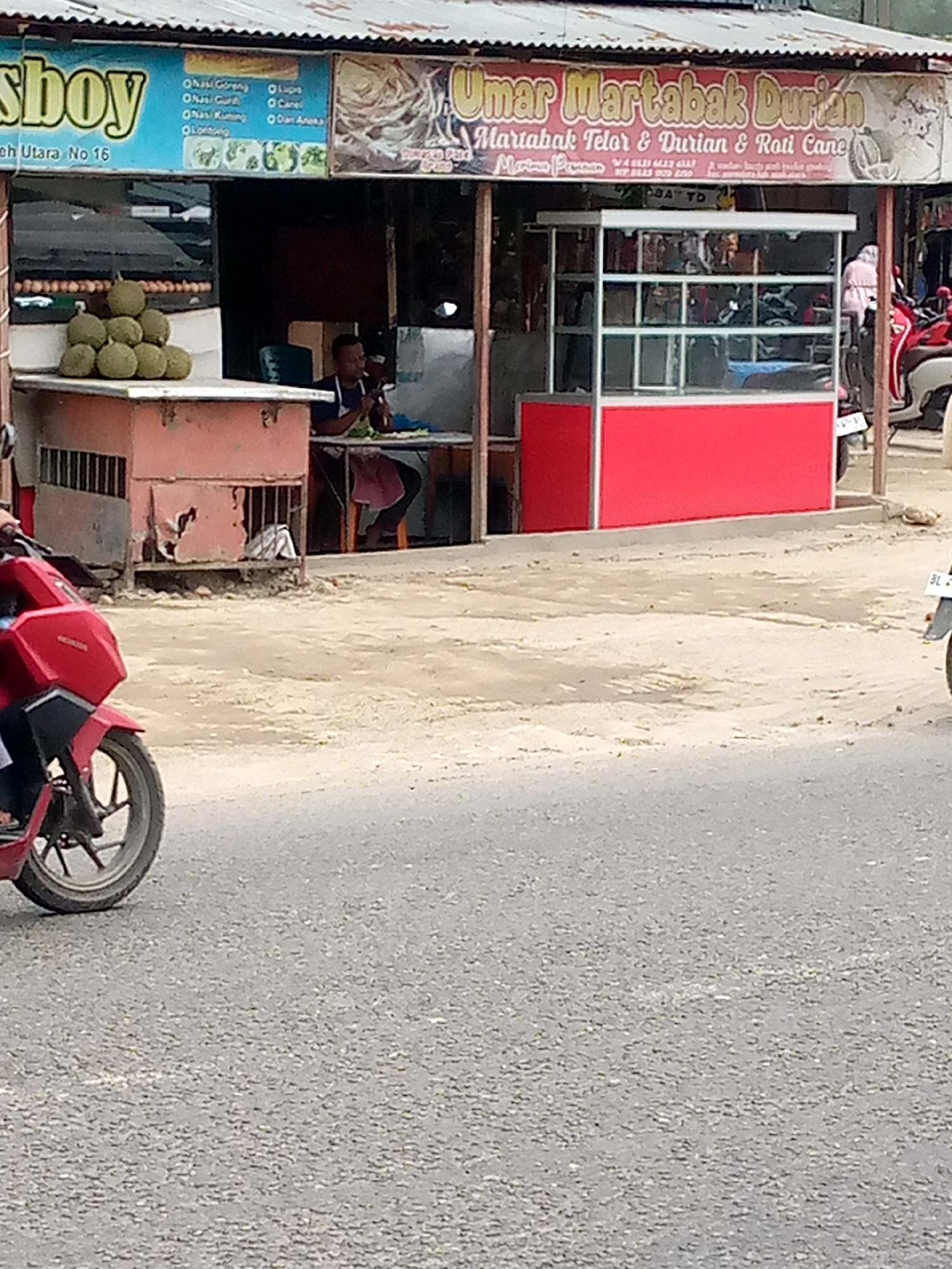 A street-level, eye-level, medium shot captures a scene in Keudegeudong, Indonesia. The focal point is a food stall with a red and white sign that reads "Umar Martabak Durian Martabak Telor & Durian & Roti Cane." Behind the counter, a person is seated, appearing to be working or eating. To the left of the stall, a pile of durians is displayed. In the foreground, the front of a red motorcycle is visible, with its wheel and part of the frame. The background shows other stalls and some parked vehicles, including motorcycles. The ground is paved with asphalt, and there's some dirt and debris on the sides. The overall impression is a busy street-side food vendor area.