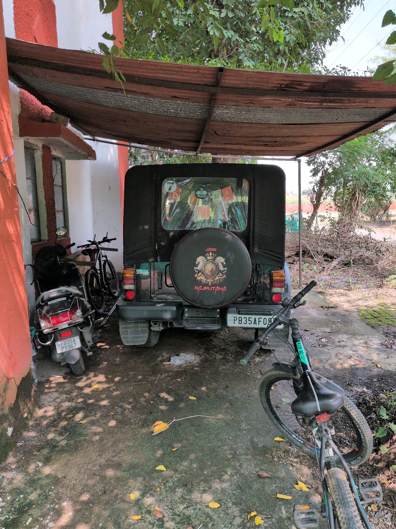 The image shows an outdoor scene under a dilapidated corrugated metal awning. In the center, a black Mahindra Thar jeep is parked. To its left, a black Honda scooter is parked. A black bicycle with green accents is parked in front of the jeep, partially obscuring it. The ground is a mix of concrete and dirt, with scattered leaves. The background features trees and dry vegetation. A building with a red and white exterior is visible to the left. The jeep has a spare tire cover with the text "JAGGI RAJPUTANA" and a crest. The scooter has a license plate that reads "PB35A P8967". The jeep's license plate reads "PB35AF091". The setting appears to be a residential or utilitarian area in Pathankot, India, given the visible license plate format. There are no people visible in the image.