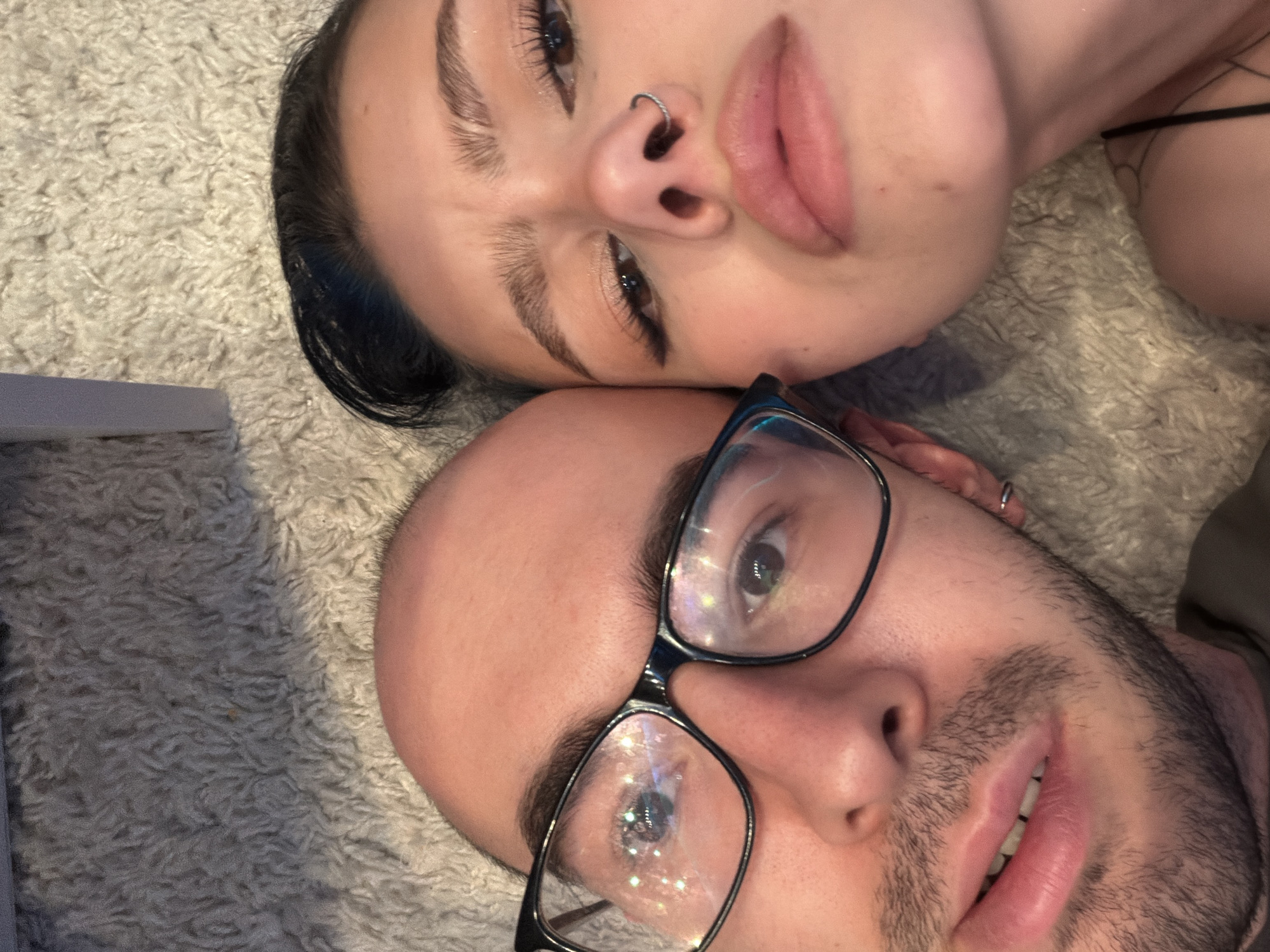 An intimate, close-up portrait shows two individuals, a female and a male, lying side-by-side on a light-colored, shaggy carpet or rug. The female is positioned above and slightly behind the male, her head resting near his. She has dark hair, a subtle nose ring in her left nostril, and her eyes are open, looking towards the camera. Her lips are slightly parted, revealing a soft, neutral expression. A black strap, possibly from a top, is visible near her shoulder.
The male is in the foreground, wearing black-framed glasses that show multiple reflections of light, creating a sparkling effect. He has short facial hair, including stubble around his mouth and chin. An earring or piercing is visible in his left ear. His lips are slightly parted in a relaxed manner, and his eyes, visible through his glasses, are also directed towards the camera. Both subjects appear comfortable and at ease, suggesting a personal and close moment. The lighting indicates an indoor setting, likely with artificial light. No text is visible in the image. The location context provided for the image is Khimki, Russia.