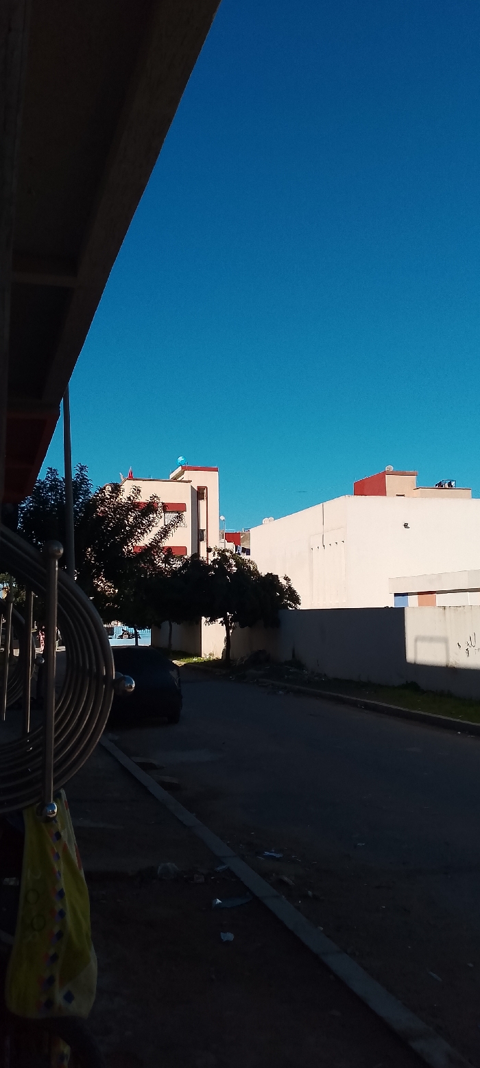 The scene depicts a street in Aïn El Aouda, Morocco, under a clear, bright blue sky. The perspective is from beneath an overhead dark structure, possibly an awning or balcony, visible in the upper left. In the immediate foreground to the left, a silver-colored metal railing or bicycle rack with spiraling elements is prominent, from which a yellow patterned cloth or bag hangs. The ground level comprises a dark, shadowed asphalt road on the right, separated by a concrete curb from a dirt verge on the left. Scattered debris, including papers and plastic, is visible on both the road and verge.
A dark-colored sedan is parked on the left side of the road, partially obscured by deep shadow. Beyond this, several trees with dark, dense foliage line the street. The midground features residential or commercial buildings. The most prominent building, slightly to the left, is light-colored (cream or off-white) with reddish-brown trim around windows and rooflines. A light blue, spherical object, possibly a water tank cover, is visible on its rooftop. Further to the right, another multi-story light-colored building with a high wall exhibits vertical indentations on its facade. Its rooftop features reddish-brown parapet walls and multiple satellite dishes or antennas. Sunlight appears to originate from the upper right, casting significant shadows across the street and foreground elements. No individuals are visible within the frame.