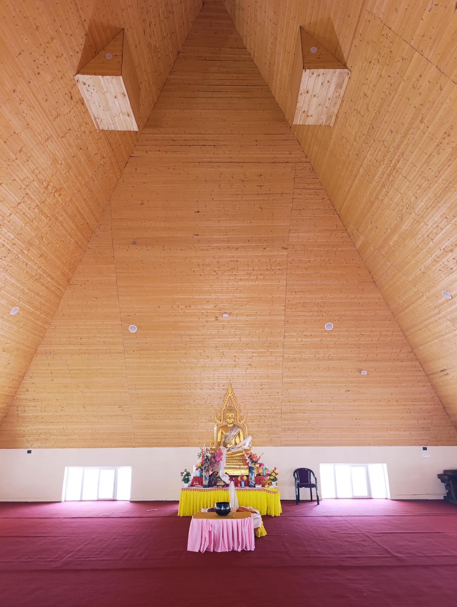 An interior view of a Buddhist temple in Thiksey, India showcases a large, triangular wooden ceiling that narrows towards the apex. Two rectangular wooden structures are inset into the ceiling on either side. A golden Buddha statue is prominently displayed on a yellow altar adorned with flowers and offerings in the center of the frame. In front of the altar, a smaller table is draped with a pink cloth. A dark chair is positioned to the right of the altar, and a red carpet covers the floor. Large windows are visible on the back wall, letting in natural light.
