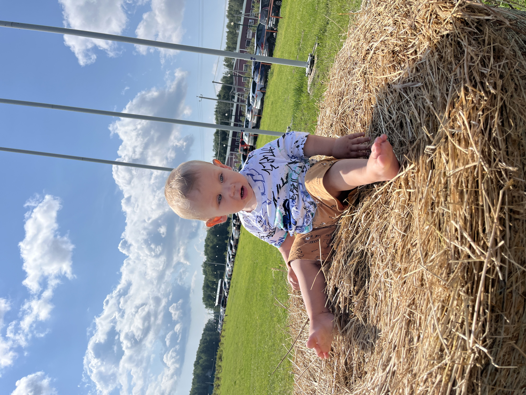 A young child with blond hair and blue eyes is sitting on a large bale of hay. The child is wearing a white t-shirt with a blue and black pattern and brown shorts with a pattern. The child is looking at the camera with a surprised expression. Their left foot is visible, and they are reaching out with their left hand towards the hay.
The setting is outdoors, with a bright blue sky filled with fluffy white clouds. A green grassy field stretches out behind the hay bale, with a line of trees in the distance. Several cars are parked in the field, and there are some buildings visible further back. There are also three vertical poles in the foreground, which are likely part of some sort of structure.
The image appears to have been taken during the daytime, with bright sunlight casting shadows. The overall mood is cheerful and playful, suggesting a fun outdoor activity. There are no visible text elements in the image. The location context provided is Akhlebaikha (Kovriginskii S/S), Russia.