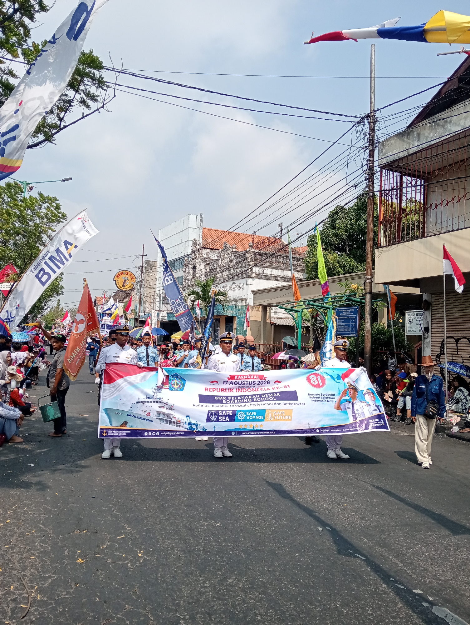 The image depicts a street parade in Bintoro, Indonesia, likely on August 17, 2026, celebrating Indonesia's 81st Independence Day. The central focus is a banner held by several individuals in uniform, promoting the SMK Pelayaran Demak (Demak Maritime Vocational School). The banner features an image of a ship, the school's emblem, and the slogans "SAFE SEA" and "SAFE FUTURE." The parade appears to be moving along a paved street lined with buildings and trees. Spectators are visible on the sides of the street, some seated and others standing. Flags and banners, including one for "BIMA," are also prominent throughout the scene. The sky is clear and bright, suggesting a daytime event.