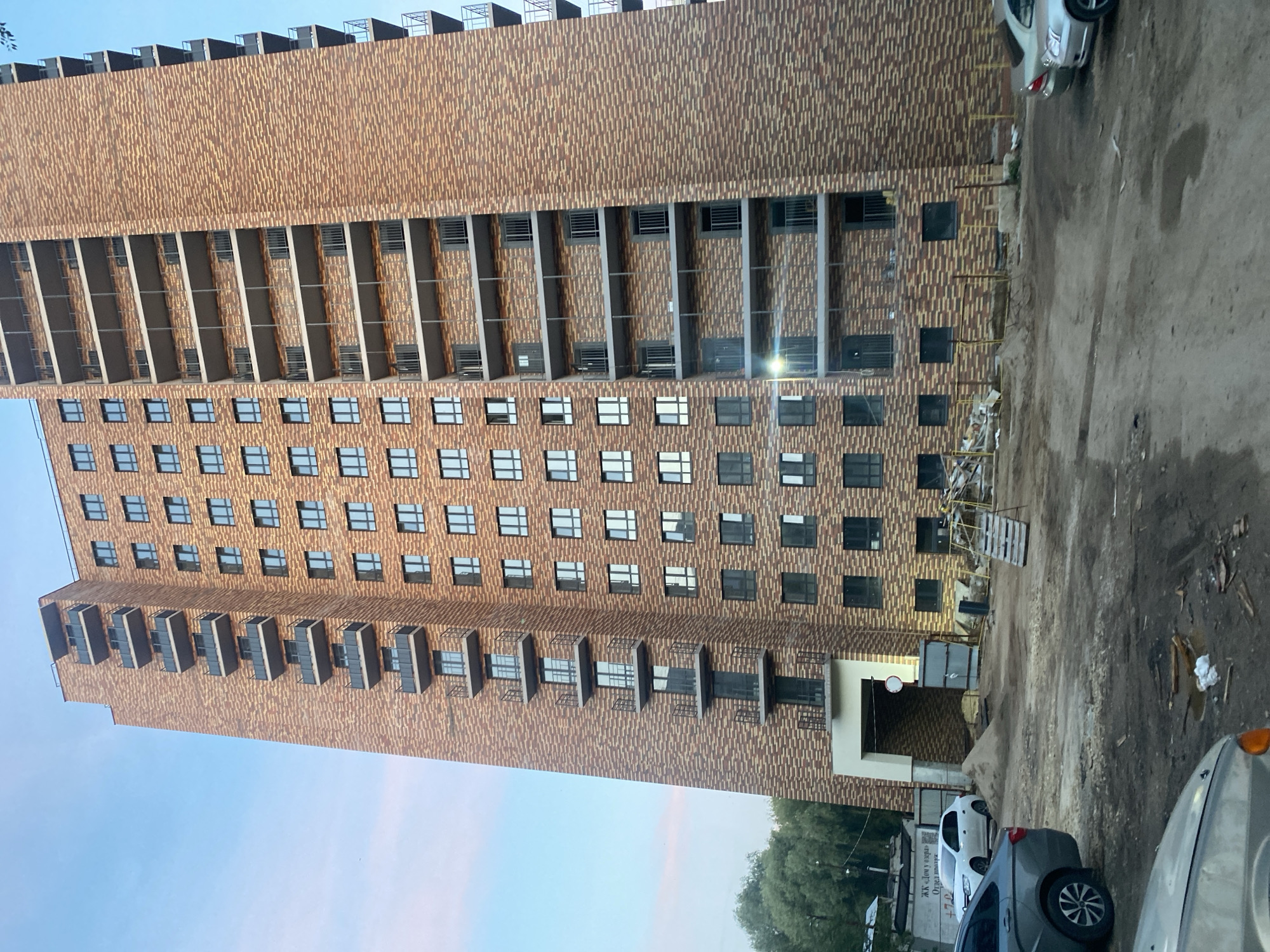 This is a low-angle, full-shot photograph of a new, multi-story brick apartment building under construction. The building is clad in light brown and dark brown bricks, with multiple balconies on each side. The sky is a clear blue, with a hint of pink on the left horizon, suggesting it is either early morning or late evening. The foreground is a muddy construction site with piles of sand, wooden planks, and construction debris. Several cars are parked in the foreground and midground, including a silver sedan, a dark gray sedan, and a white sedan parked near the building's entrance. The building entrance is a large, arched opening. There are no people visible in the image. The image appears to have been taken in the city of Samara, Russia, based on the text on a white sign to the left of the building entrance which reads "ЖК «Дом у озера»" (Residential Complex "House by the Lake") and a phone number.