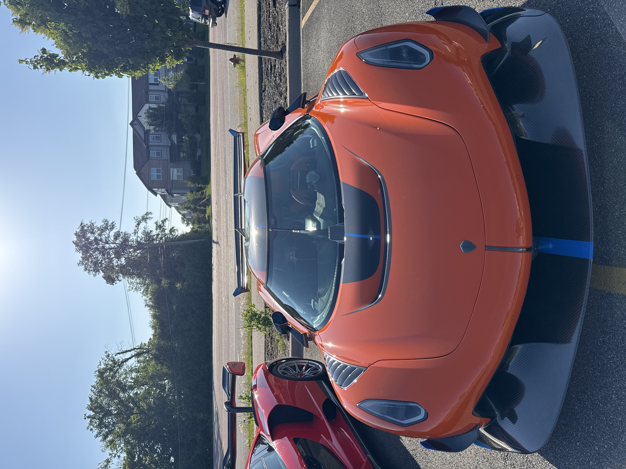 The image presents a partially obscured view of a public parking lot in Warrenville, Illinois, United States. The foreground is dominated by two high-end sports cars, parked adjacent to each other. The foremost car is an orange coupe with carbon fiber detailing, while a red coupe is partially visible behind it. The lighting indicates it is daytime, and shadows suggest the sun is positioned relatively high in the sky. The surface of the parking lot appears to be asphalt.
In the background, a line of trees partially obstructs a residential building, likely an apartment complex. There is also a utility pole and another vehicle parked further away. The sky is predominantly clear and blue. No people are visible.