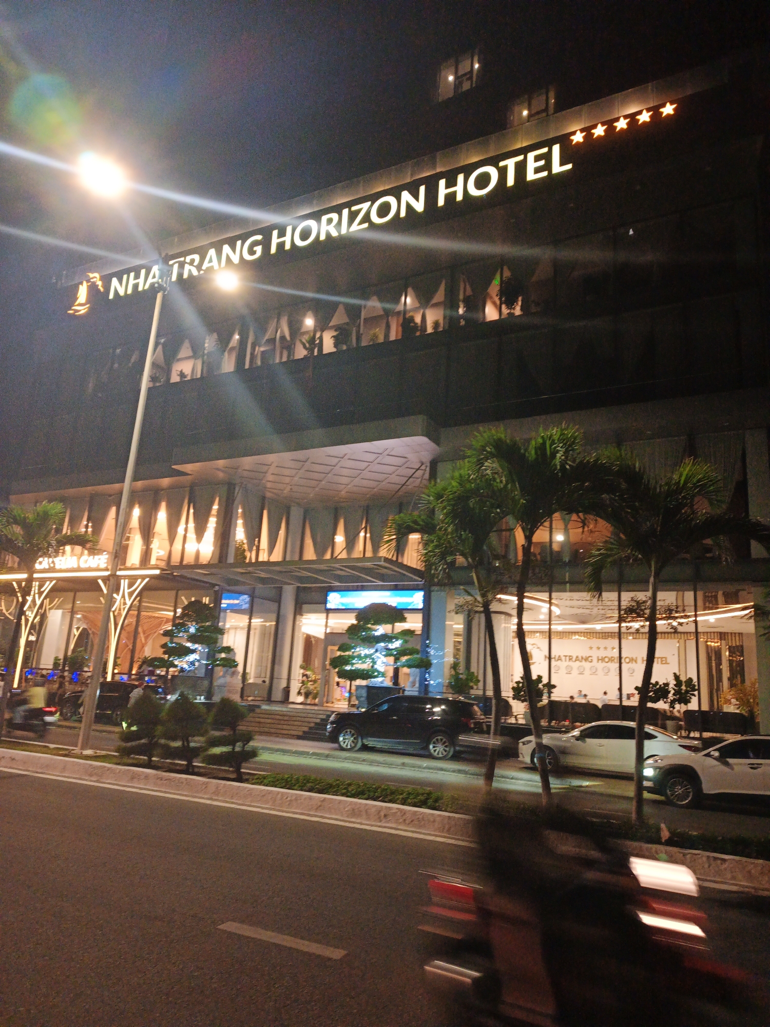 The image depicts a nighttime scene in Bac Nha Trang Ward, Vietnam, focusing on the illuminated facade of the "NHA TRANG HORIZON HOTEL". The hotel's name is prominently displayed in large, lit letters across the top of the building, accompanied by five orange stars.
The hotel itself is a modern structure with multiple levels, featuring large windows that reveal interior lighting. Decorative elements, including stylized trees and palm trees, are strategically placed around the entrance and along the sidewalk. The entrance area is lit by warm, ambient lights, and a sign for "CAFE" is visible.
In front of the hotel, cars are parked along the street, including a white sedan and a dark SUV. A motorcycle with a blurry rider is captured in motion on the road, indicating a busy street even at night. The road surface is asphalt, with white lane markings and a concrete curb.
The time of day is clearly night, with the sky being dark and the artificial lights of the hotel and street illuminating the scene. The weather appears clear, with no visible rain or fog. The overall atmosphere is one of urban activity and commercial presence, characteristic of a well-lit hotel district at night. The image is captured from a street-level perspective, looking up at the hotel.
