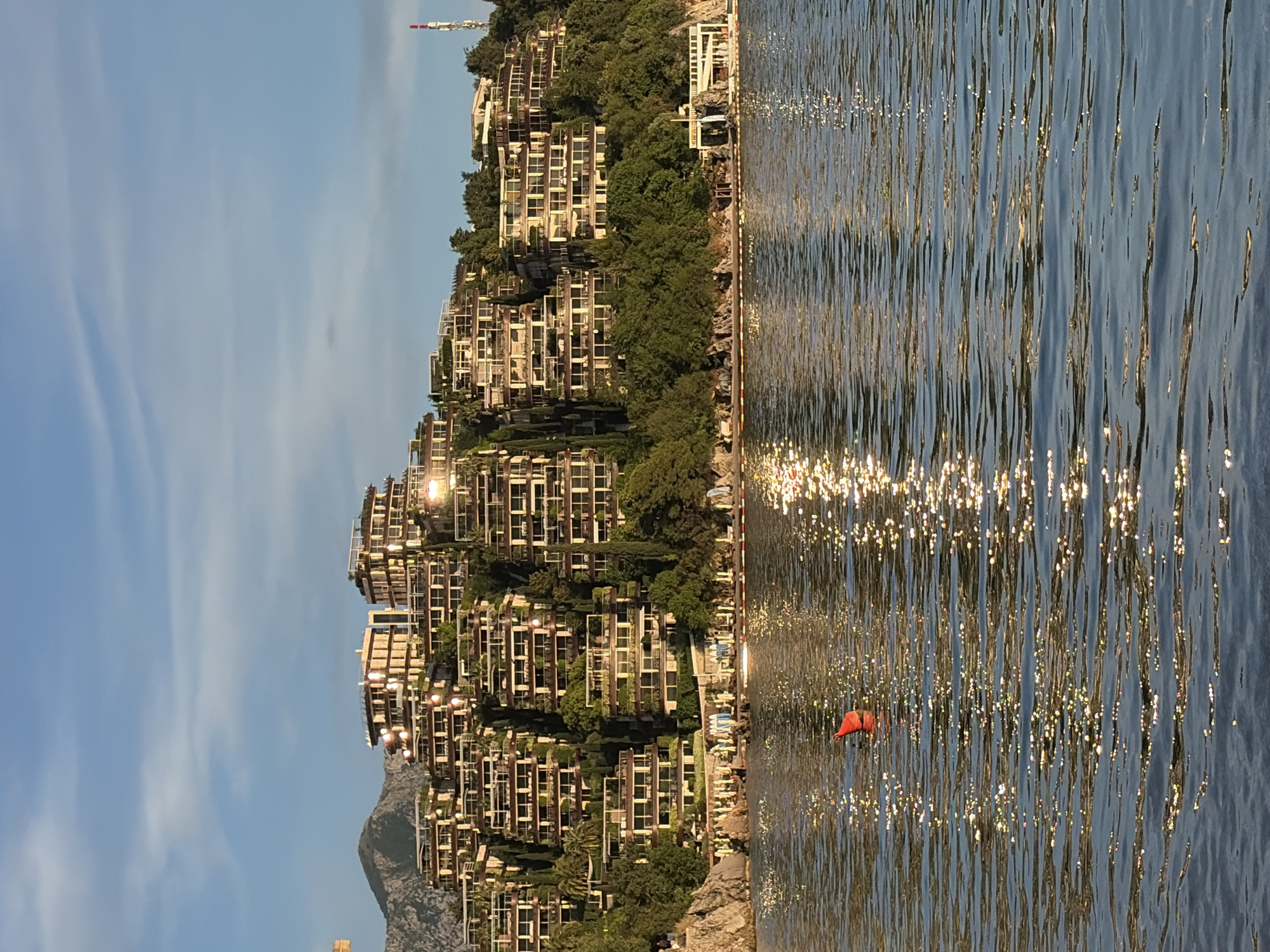 The image shows a shoreline scene in Budva, Montenegro, featuring tiered buildings nestled into a hillside covered with trees. The Adriatic Sea shimmers with reflected sunlight. An orange buoy floats in the water near the shore. The sky is clear with light cloud streaks.
