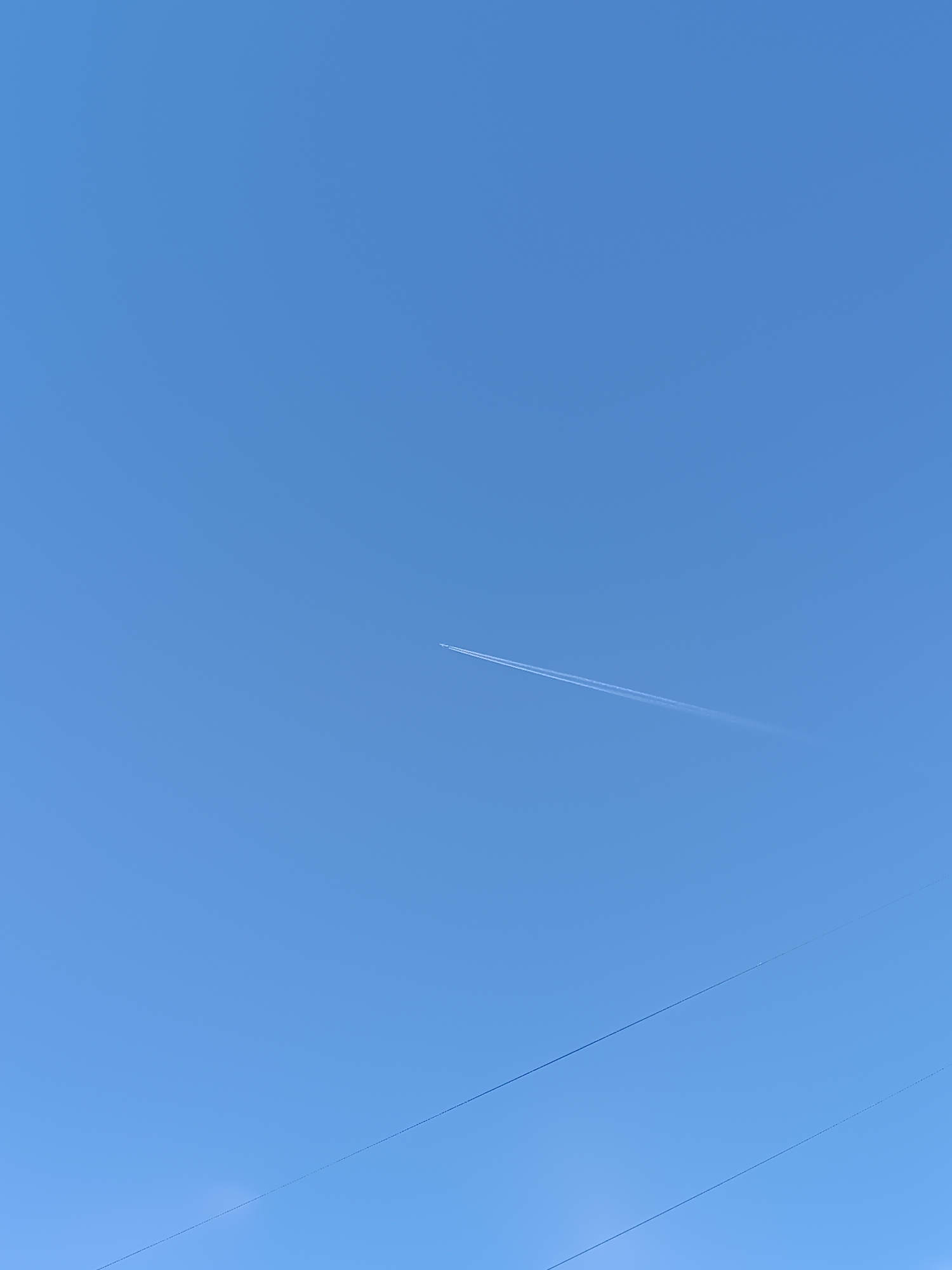 A clear blue sky dominates the scene. A distant airplane with a visible contrail flies diagonally across the frame, from the upper-left towards the lower-right. In the lower portion of the image, three horizontal, dark power lines stretch across the frame. The location and whether the photograph was taken in Chaikovsky, Russia, are unknown.