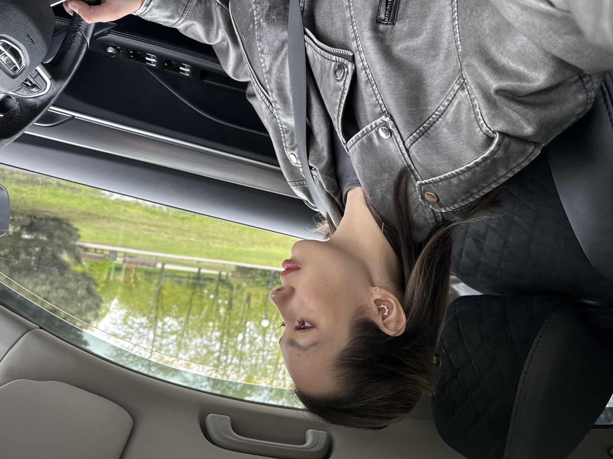 A woman is in a car. She is wearing a gray jacket and a seatbelt. Her right ear is visible, adorned with a crescent moon earring and another dangling earring. Outside the car window is a view of a pond with greenery. The location is in the city of Unknown, Unknown.