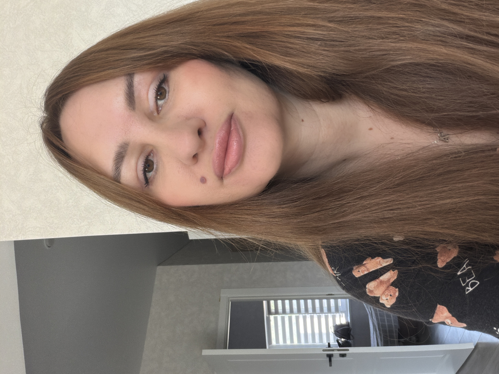 The image shows a close-up, head-and-shoulders portrait of a woman with long, auburn hair. She has fair skin, brown eyes, and is wearing light makeup, including lipstick and eyeliner. A mole is visible on her left cheekbone, and another small mole on her neck. She is wearing a dark, long-sleeved shirt with a pattern of teddy bears and the word "BEAR" visible.
The background is slightly blurred but shows an interior setting. To the left, a white door with a silver handle is ajar, revealing a glimpse of a room with a window covered by blinds. The walls appear to be a light beige color. The overall impression is that this is a casual, indoor photograph, likely taken in a residential environment. No specific details point to Malinovka, Russia.