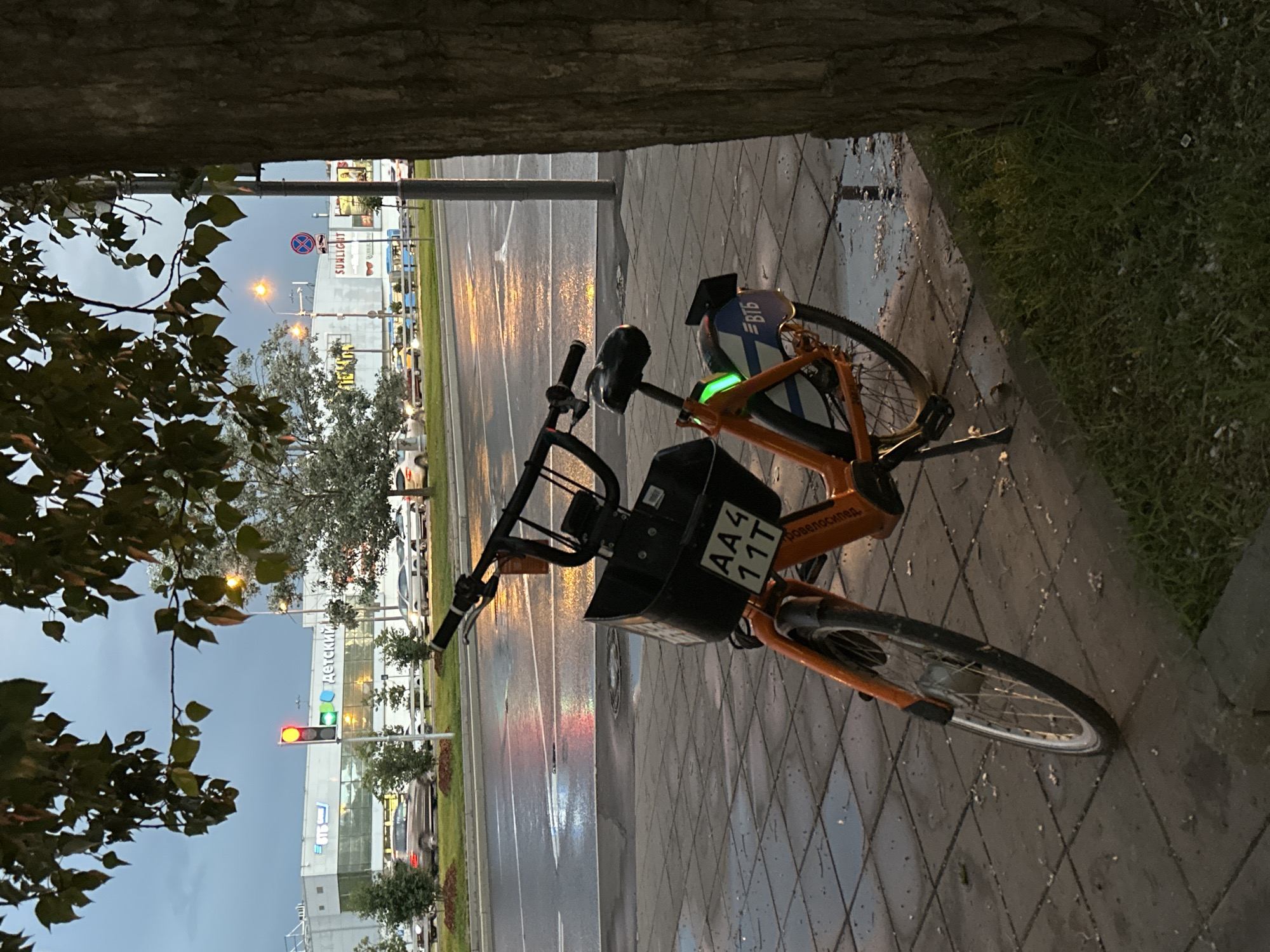 An orange public bike is parked on a wet sidewalk next to a tree on a cloudy evening in Moscow, Russia. The bike has a basket in front and a license plate that reads "AA4 11T". The street behind the bike is wet from rain, reflecting the lights of nearby buildings. In the background, there are several stores and a traffic light that is red and green. The sky is overcast and the overall mood is calm and somber.